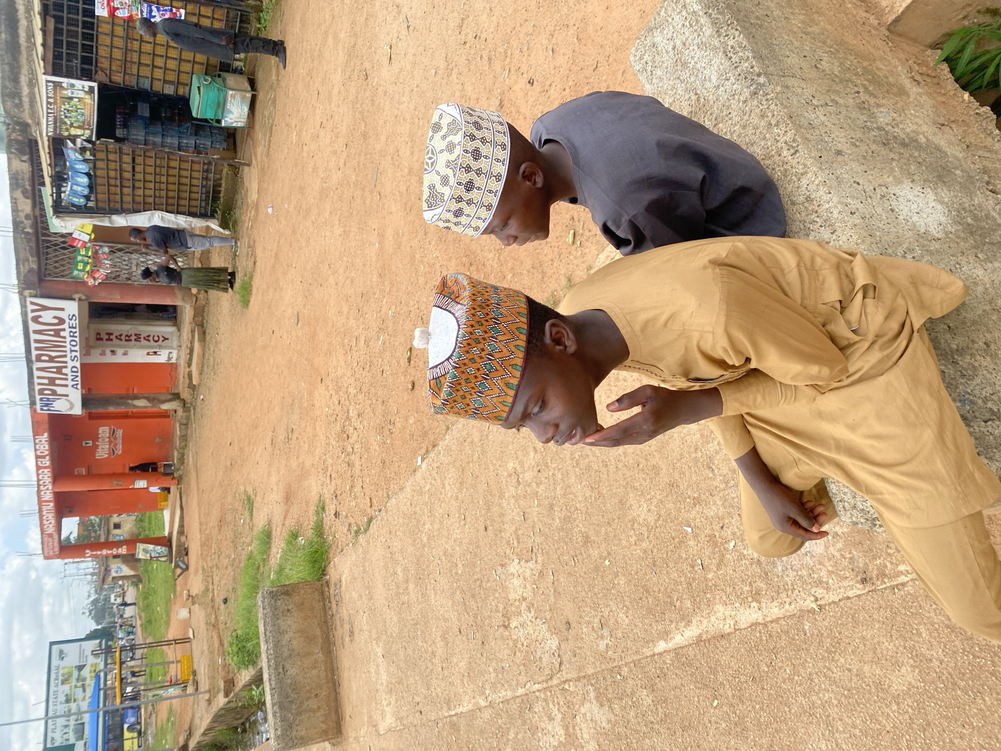 In Bukuru, Nigeria, two young boys sit facing right, on a concrete barrier with a sandy, reddish-brown dirt ground behind them. The boy on the left is wearing a tan, long-sleeved tunic and matching pants, topped with an ornate, patterned cap. He is resting his chin on his left hand, looking thoughtfully to the right. The boy next to him, on the right, wears a dark grey tunic and a white and gold patterned cap. He is also looking to the right.
In the background, there are several buildings, one brightly colored orange with signage that reads "NASAMU NASARA GLOBAL" and "FNR PHARMACY AND STORES." Another building, to the right, has a metal grate facade and signage reading "NW ANMA F.C & SONS". A few people are visible interacting near these shops. A bus is parked to the far left, with a billboard behind it. The sky is overcast.