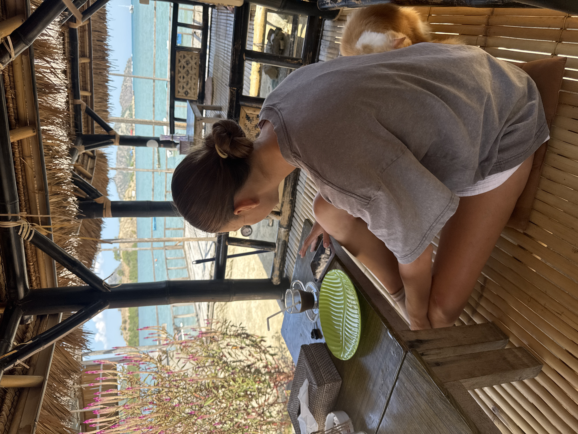 A woman with her hair pulled back in a bun sits cross-legged on a bamboo floor in an open-air pavilion. She is wearing a grey t-shirt and white shorts. In front of her on a wooden table is a cup of coffee with a straw, a green leaf-shaped plate, a tissue box, and a smartphone. Her left hand is resting on the phone. To her right, a ginger and white cat is visible. Through the open sides of the pavilion, a view of the ocean, a wooden pier, and a distant shoreline with hills can be seen. The pavilion has a thatched roof and bamboo supports. The scene appears to be located in Kertasari, Indonesia, likely at a beachfront establishment.