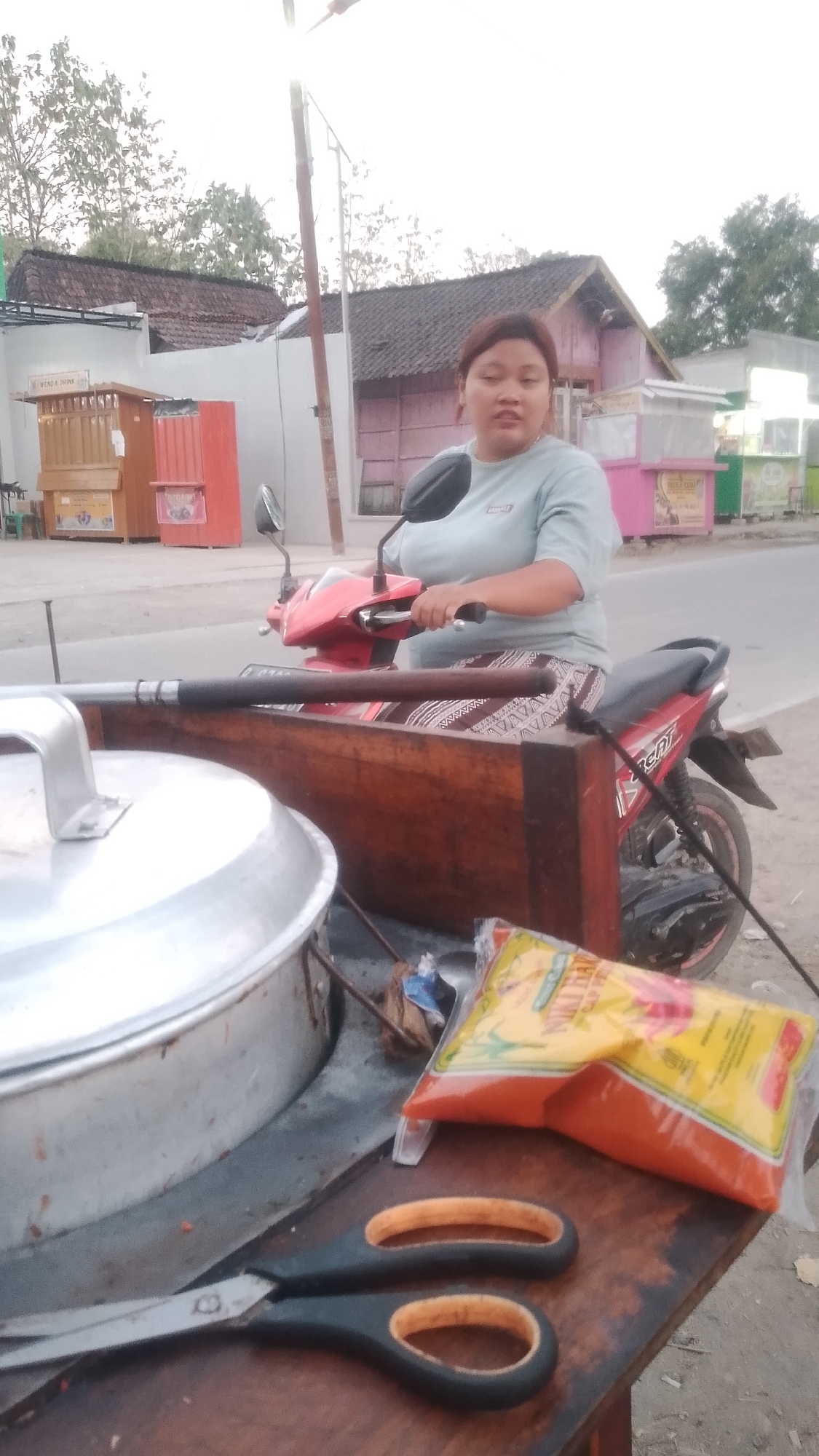 In the city of Karanglangu, Indonesia, a woman with dark hair is seated on a red and black scooter. She wears a light blue t-shirt and a patterned skirt. In front of her is a wooden cart. On the cart are a large silver pot with a lid, a pair of black scissors with orange handles, and a sealed plastic bag containing an orange substance. Behind the woman, buildings with tiled roofs line the street. The sky is bright, suggesting daylight.