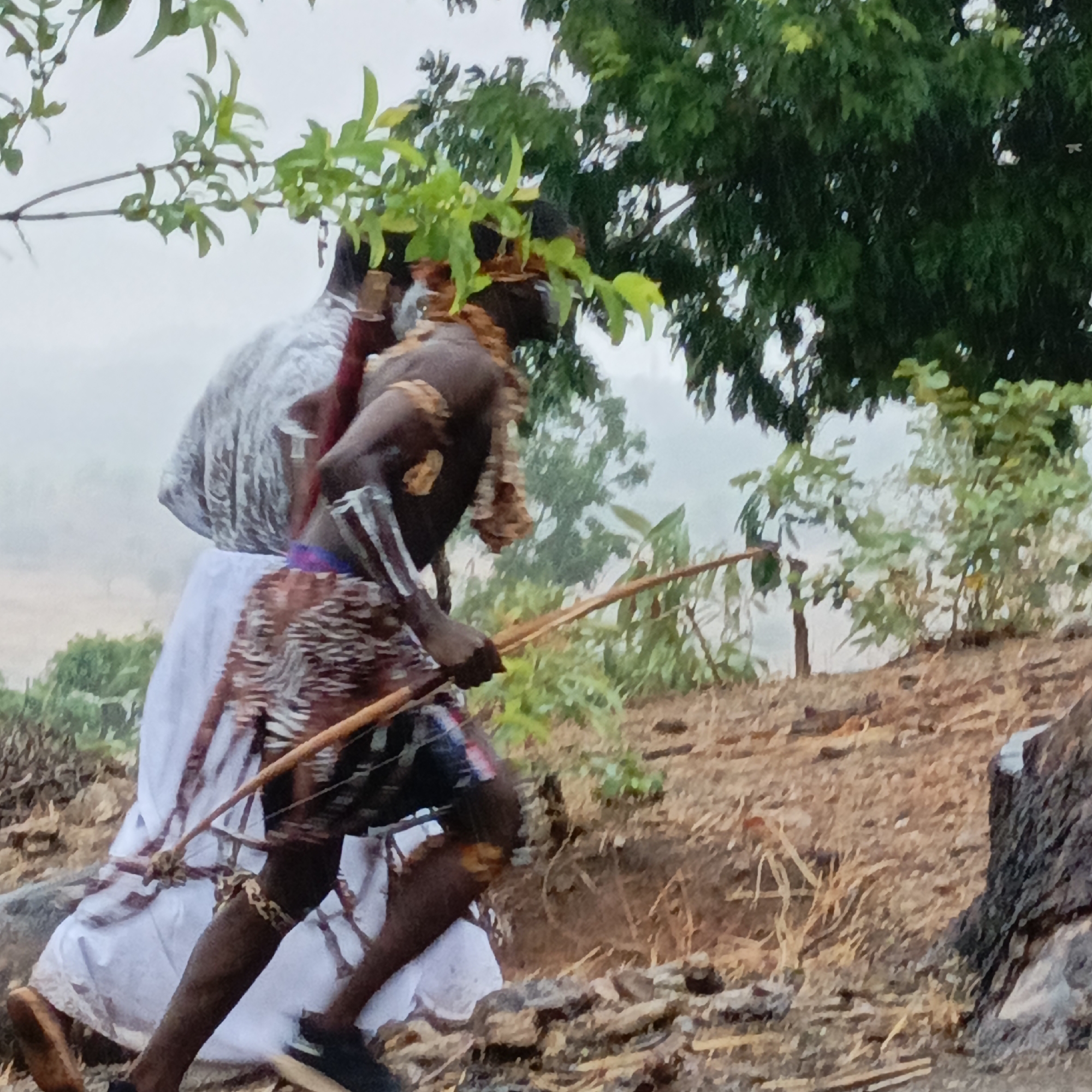 In an outdoor setting in Boher, Nigeria, two dark-skinned individuals are captured in action. The foremost figure is a man in mid-stride, holding a bow, with what appears to be a quiver of arrows secured to his back. Adornments include plant material on his head and around his neck, and a bracelet on his leg. The second person is draped in a white cloth. The environment consists of a slightly hilly terrain with dry vegetation and trees in the background, suggesting a rural or natural area. The lighting is subdued, potentially due to weather conditions or time of day.