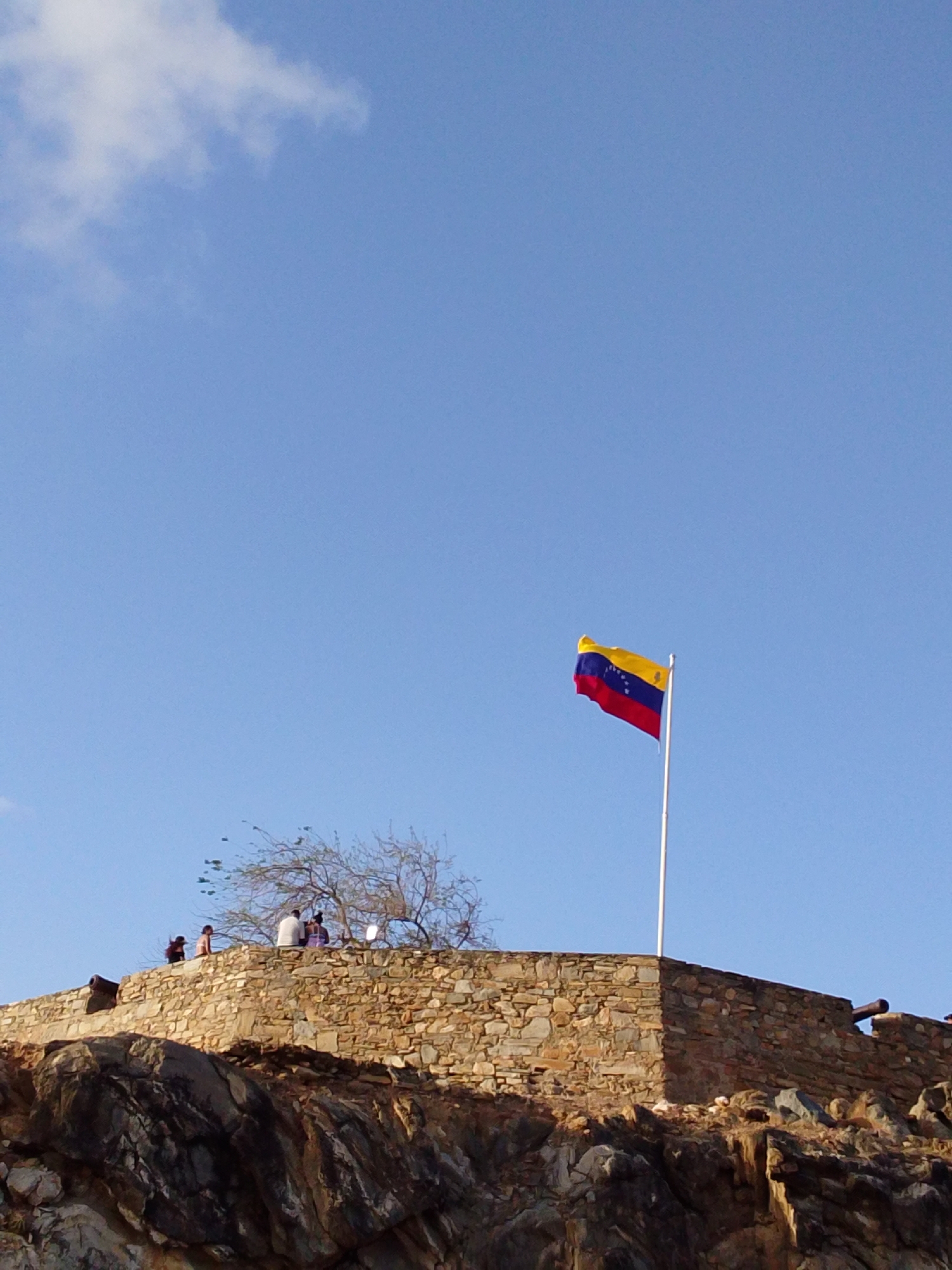 The image displays a historical fort or defensive structure situated on a rugged, rocky elevation beneath a clear, bright blue sky with sparse white clouds. Dominating the upper right portion of the frame is a vibrant Venezuelan flag, featuring horizontal yellow, blue, and red stripes with an arc of white stars on the blue band, flying from a tall flagpole.
The fort itself is constructed from large, irregular stone blocks, forming a robust wall along the cliff's edge. On top of this wall, several people are gathered. They appear to be casually observing their surroundings or resting, their figures silhouetted or dimly lit. Near the group of people, a tree with bare branches stands, its delicate structure contrasting with the solid stone. Two old cannons are visible along the fort's walls—one on the far left and another on the far right—serving as remnants of its historical military purpose.
The lighting suggests a pleasant time of day, likely late afternoon or early morning, with a warm, soft glow illuminating the stone and the people. The weather appears clear and sunny. The scene evokes a sense of history and observation. No visible text is present in the image.