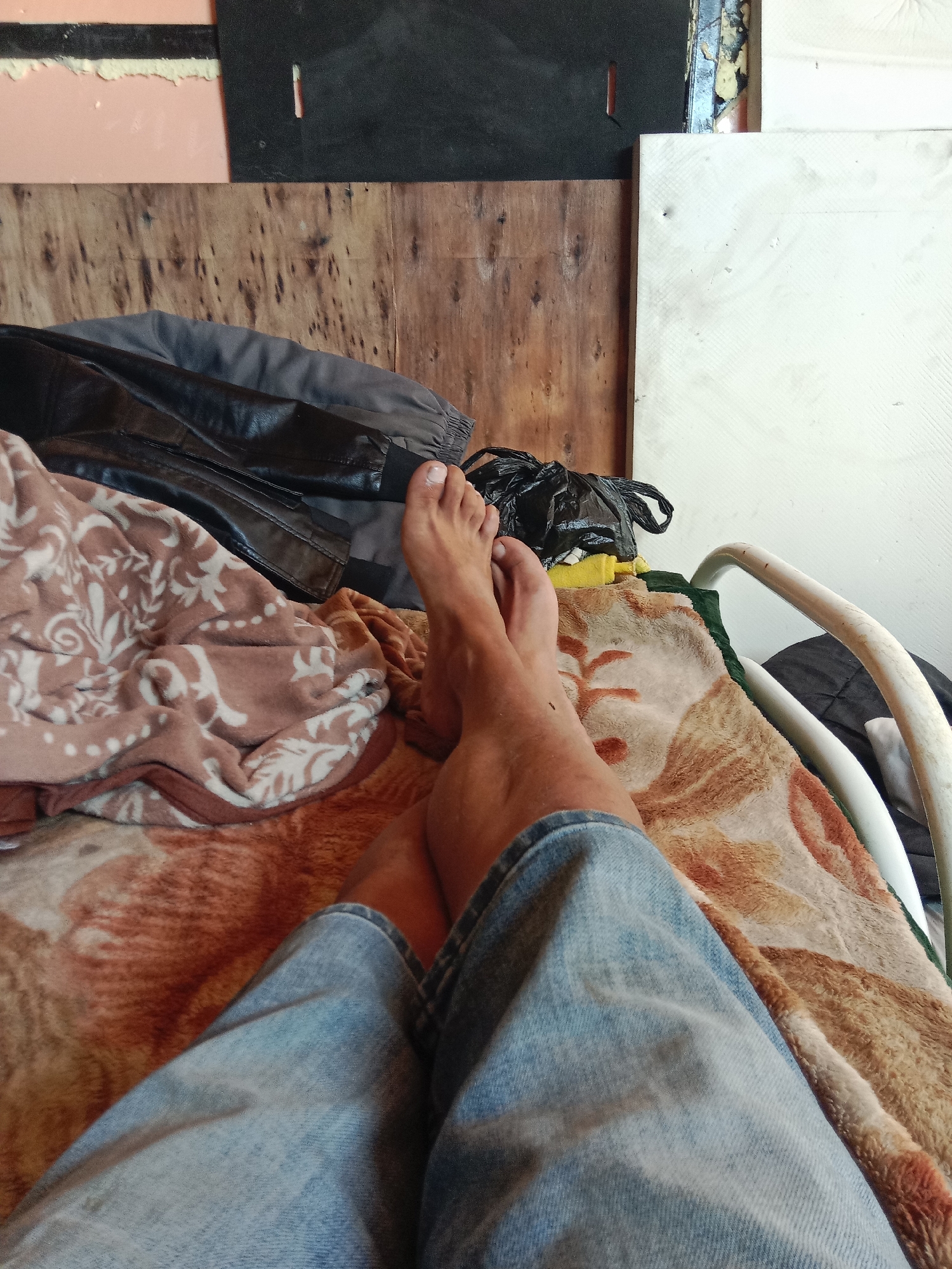 The image captures a close-up, low-angle perspective of a person's legs and feet, wearing blue denim jeans and resting on a patterned blanket. The background consists of various materials, including wooden planks, a black material, and a white textured surface. A portion of a metal railing is visible on the right side. There are also several dark-colored bags or bundles in the background, one of which is a black plastic bag. The scene appears to be indoors, possibly in a makeshift or rustic setting. There is no indication of the city of Akzhar, Kazakhstan, in the image itself.