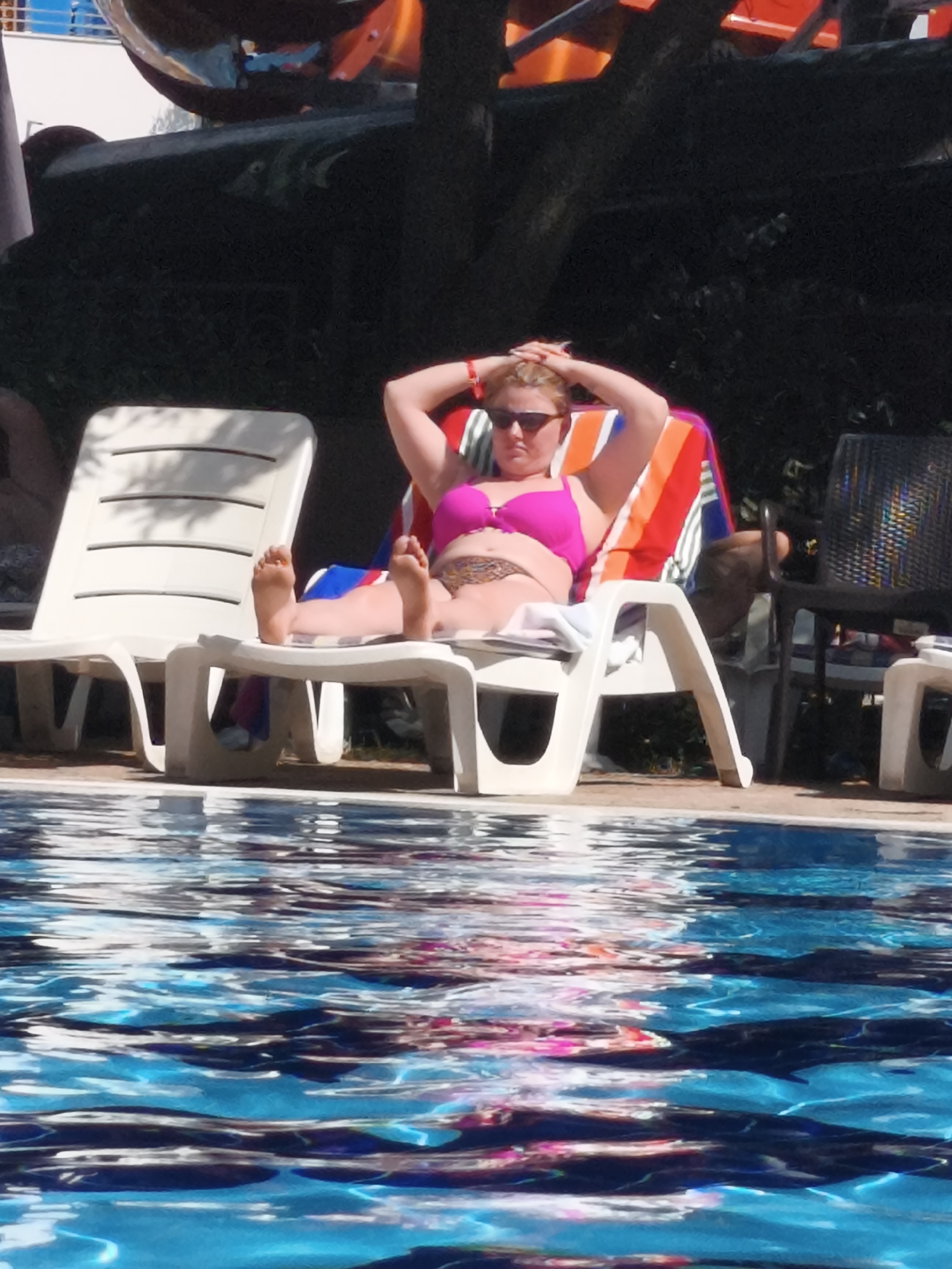 A woman is lying on a lounge chair near a pool, basking in the sun. She is wearing sunglasses and a pink bikini. The water in the pool is reflecting the sunlight, creating a shimmering effect. Behind the woman, there is a lounge chair and some greenery. In the background, there is a slide. The scene appears to be a resort or a public pool.