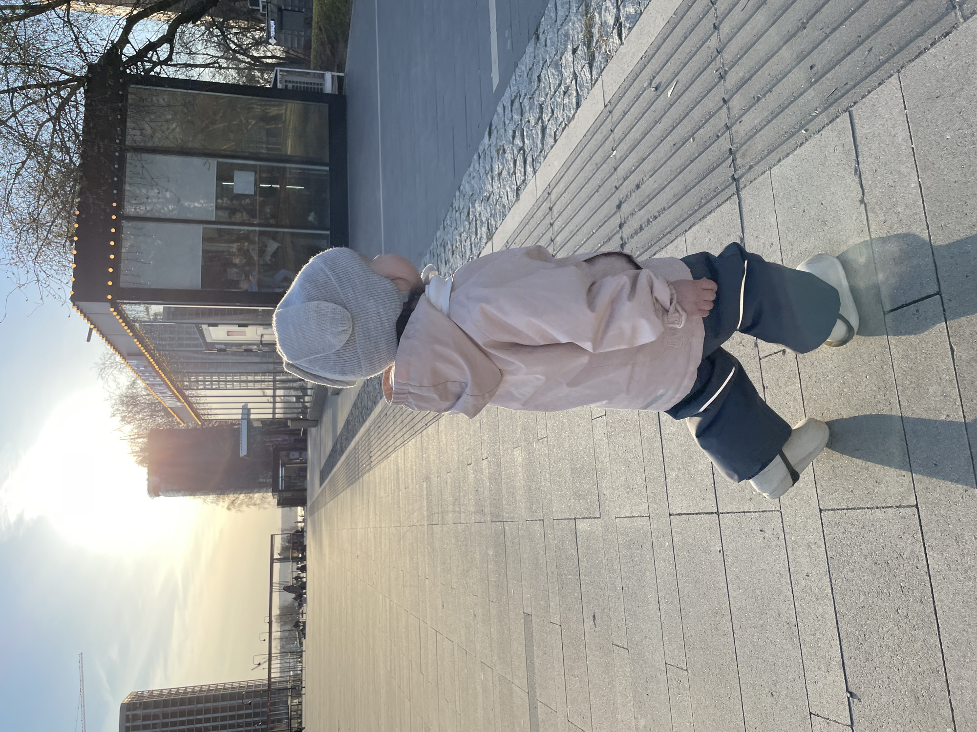 A child wearing a light-colored jacket and hat is walking on a paved sidewalk in Izhevsk, Russia. The sidewalk is made of rectangular gray stone blocks. The child is walking away from the viewer towards a bright, low sun. In the background, there is a row of trees and a building with large windows. A building with lights is visible to the left. In the distance, there are tall buildings and a cloudy sky.