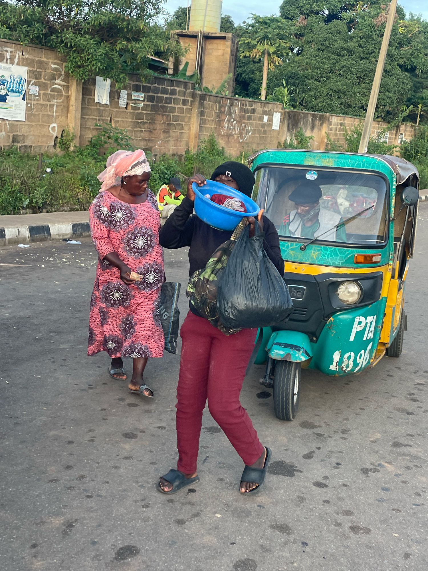 A woman in a pink dress and headscarf stands on a paved road, looking down at a plastic bowl of clothes held by another person. The person holding the bowl wears a black hooded sweatshirt and dark red pants, and carries a black plastic bag. They are positioned near a teal and yellow auto-rickshaw, which has the numbers "1896" and "PTA" painted on its side. The rickshaw has a driver visible inside. In the background, a brick wall and lush green foliage suggest an outdoor setting. The scene appears to be a street in Jos, Nigeria, likely during daylight hours given the lighting.