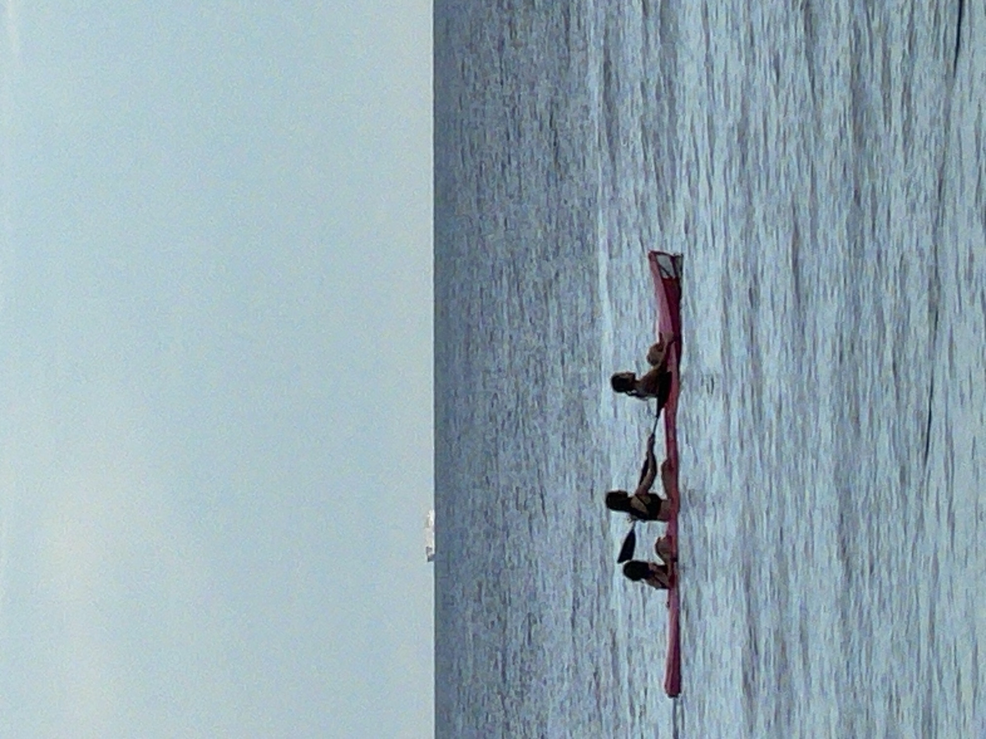 Three individuals are in a red kayak on a body of water. The kayak is positioned horizontally in the frame, with the individuals facing towards the left. The person in the front left appears to be a child, with their back to the camera. The person in the middle is an adult, also facing left, holding a paddle and appearing to row. The third person, in the rear right, is partially visible and also facing left. The water is blue and appears slightly choppy. In the distance, a large ship or barge is visible on the horizon, situated between the water and a pale blue sky. The scene is set outdoors, likely in the ocean, with no visible urban structures or city landmarks of Dauin, Philippines.