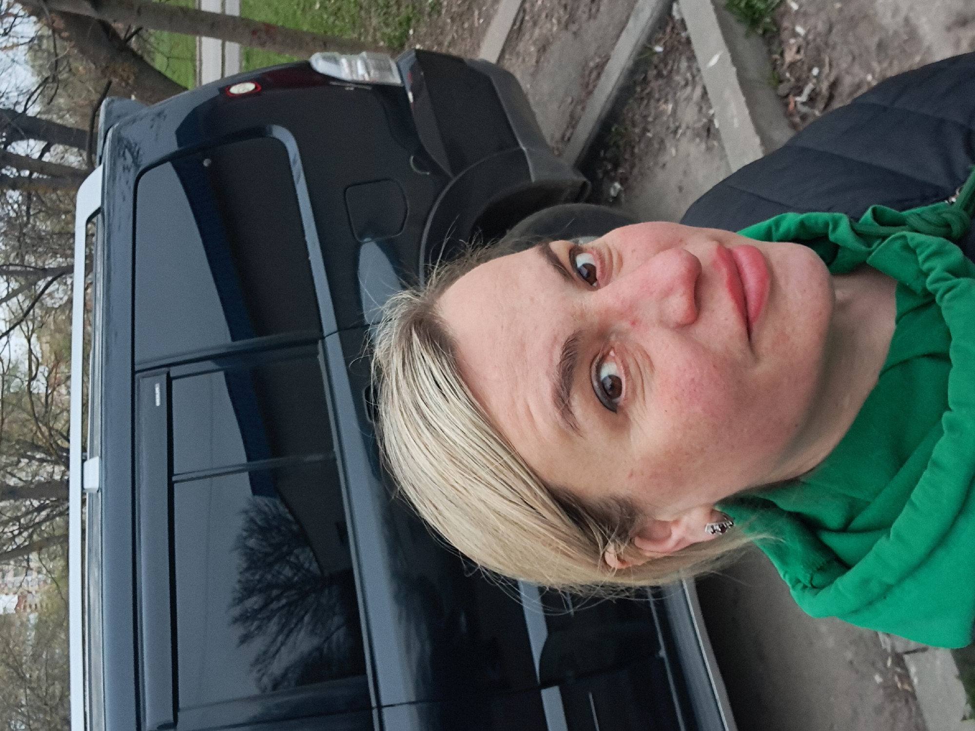 The image is a rotated, close-up, selfie-style photograph taken outdoors in Elektrostal, Russia. It features a woman in the foreground looking directly at the camera, with a dark-colored van and natural elements in the background.
The woman has blonde hair, fair skin with some redness, and wide-open brown eyes. Her lips are slightly parted. She is wearing a bright green hooded sweatshirt, with the hood pulled up but not covering her head, underneath a dark-colored jacket. A small, shiny earring is visible in her left ear. Her expression appears curious or neutral, with a direct gaze.
Behind the woman, a dark blue or black van or minivan is parked. Part of its rear and side are visible, including tinted windows and a section of a taillight. A white trim piece runs vertically along the side of the van. The scene is set on what appears to be a street or parking area, with a concrete curb separating it from an unpaved area with patches of dried leaves, grass, and dirt. Bare tree branches, characteristic of late fall, winter, or early spring, are visible through and above the vehicle.
The lighting suggests it is daytime, possibly overcast, given the soft, diffuse light and lack of harsh shadows. The image is oriented sideways, appearing rotated 90 degrees counter-clockwise from a typical portrait capture. No clear, readable text is visible in the image.