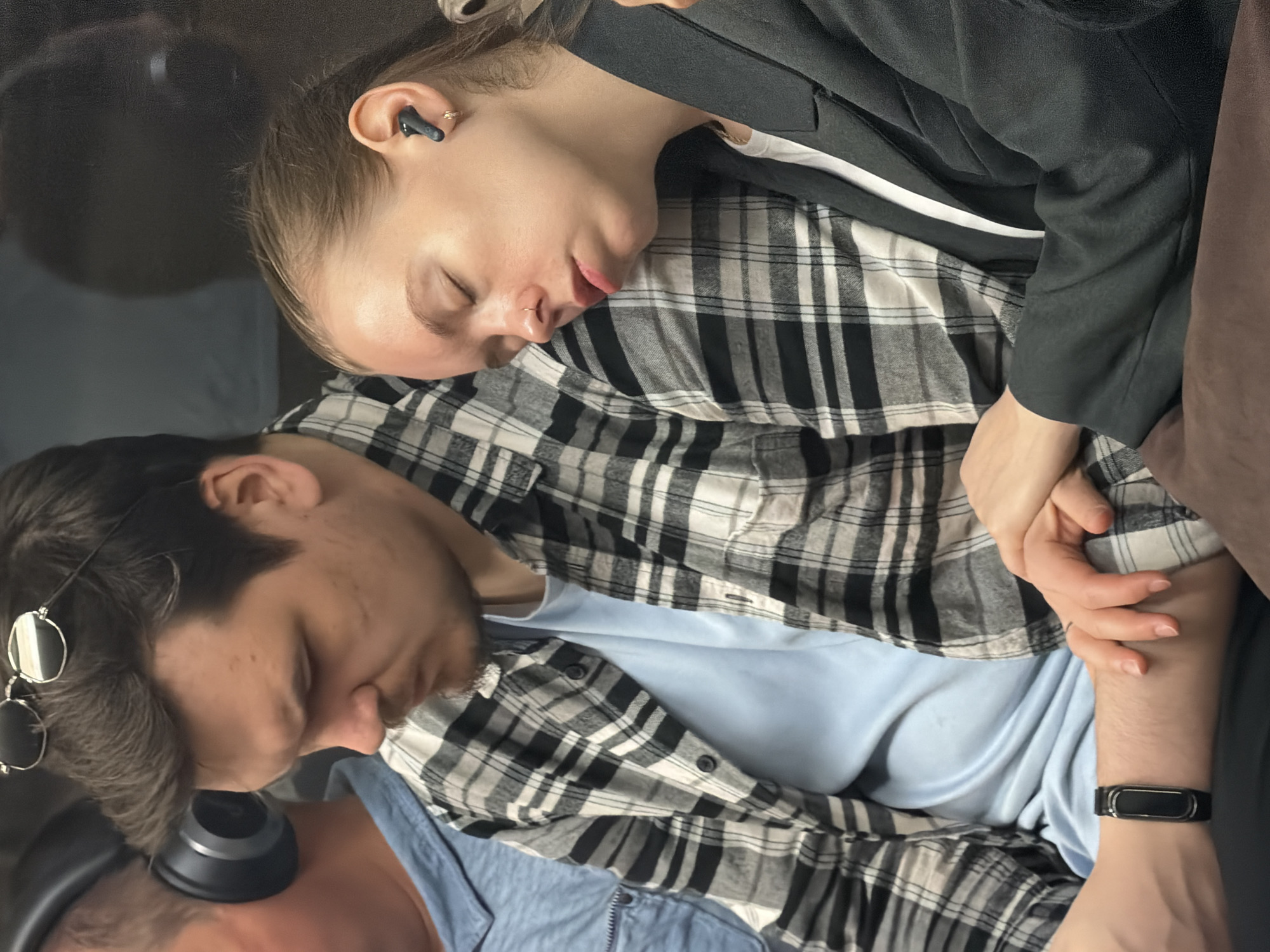 The image shows two individuals, a man and a woman, sleeping on public transportation, likely a train or bus, within the city of Moscow, Russia. The man, wearing a plaid shirt and a light blue t-shirt, is in the foreground on the left, with headphones on his head and a fitness tracker on his wrist. He appears to be looking down. The woman, to his right, is leaning her head on his shoulder, her eyes closed, wearing a black blazer over a white top and an earbud in her right ear. Her left hand is resting on the man's forearm. The environment appears to be the interior of a vehicle, with muted lighting and a reflective surface in the background.