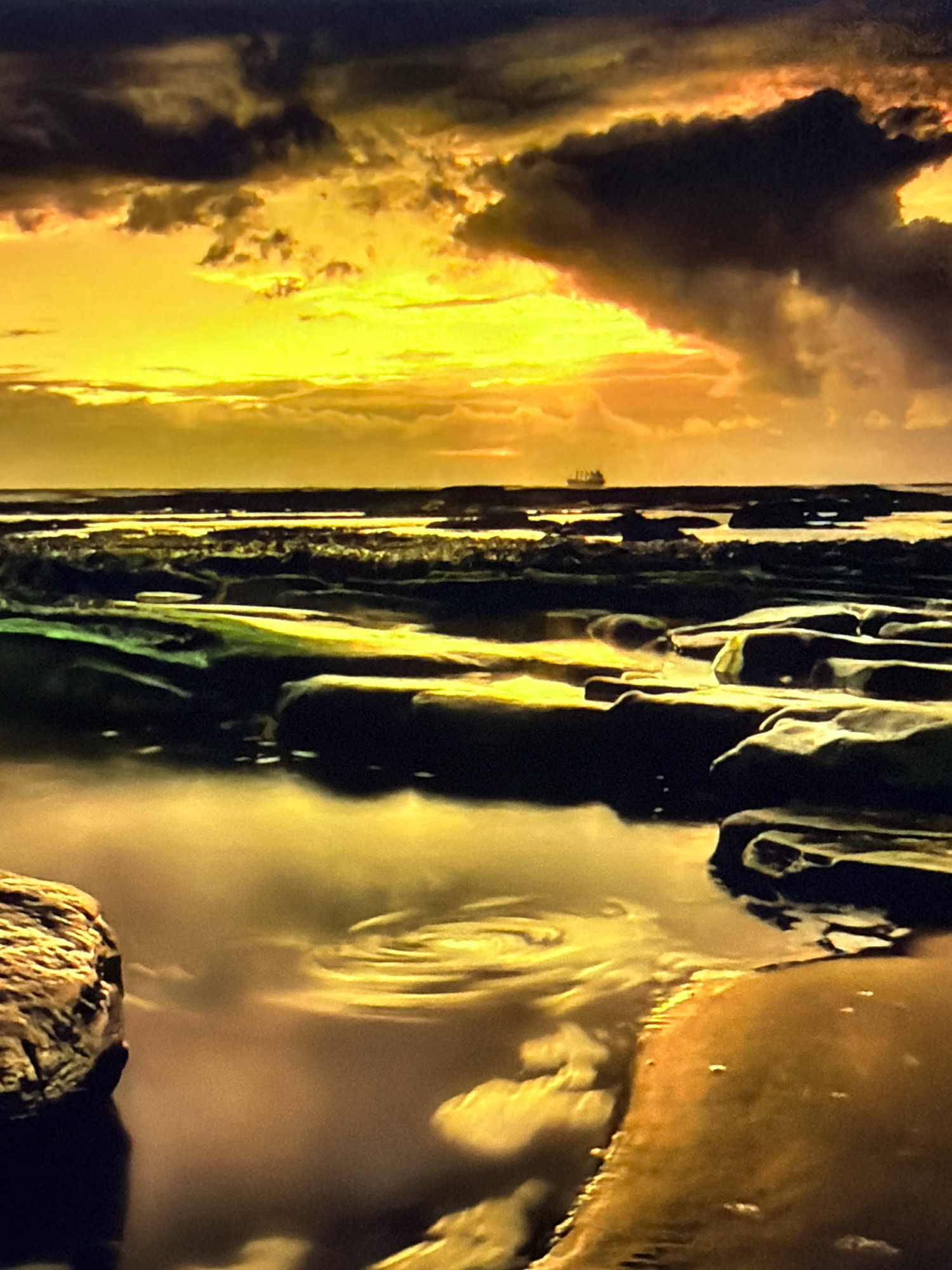 The image depicts a dramatic sunset over a rocky coastline. The sky is filled with intense yellow and orange light, broken by dark, ominous clouds. In the distance, a solitary ship is visible on the horizon. The foreground is dominated by dark, wet rocks and shallow pools of water reflecting the vibrant sky. The overall impression is one of raw, natural beauty and a sense of dramatic weather. There are no people or explicit signs of human activity in the image.
Kafr Yasif is a town located in the Northern District of Israel.