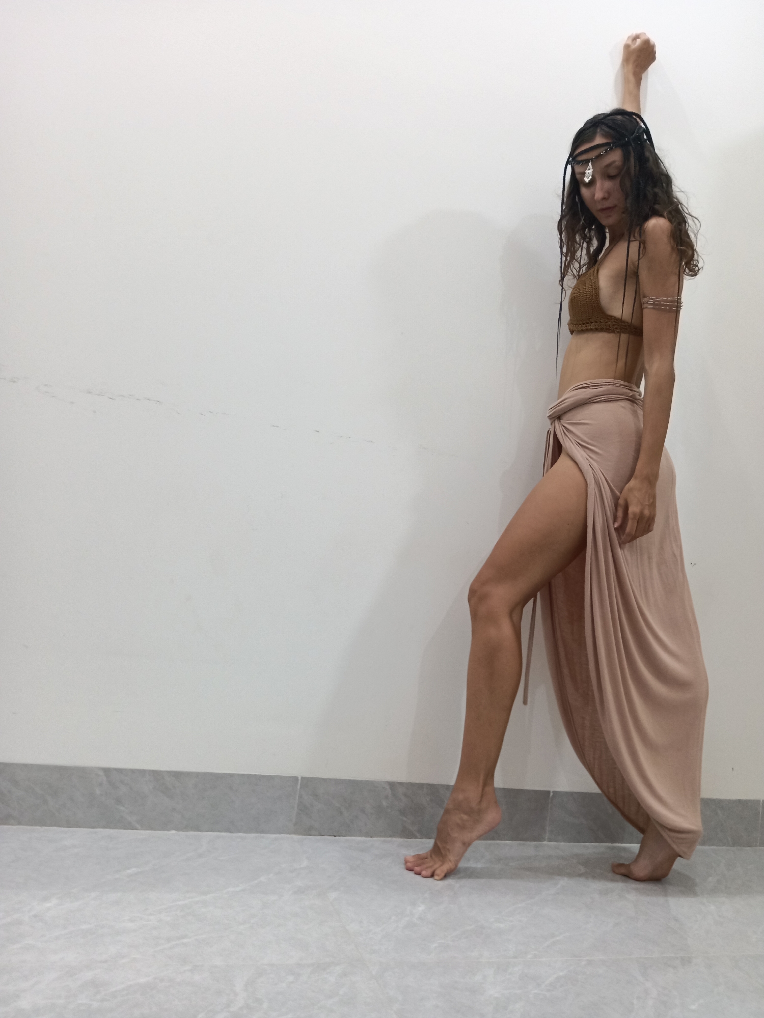 A young woman stands in a room with a white wall and a tiled floor. She is leaning against the wall with her right arm raised and her hand resting on the wall. She is wearing a brown crocheted bikini top, a light-colored wrap skirt with a high slit, and a headpiece. Her hair is long and curly, with braided strands adorned with dark cords. She is also wearing armbands on her upper arms. Her left leg is bent at the knee, with her foot resting on its toes, while her right leg is extended behind her, with her foot on the floor. The scene appears to be a dance or modeling pose, and the lighting suggests an indoor setting, possibly in Mũi Né, Vietnam, given the style of dress and overall aesthetic that might be associated with a coastal resort town.