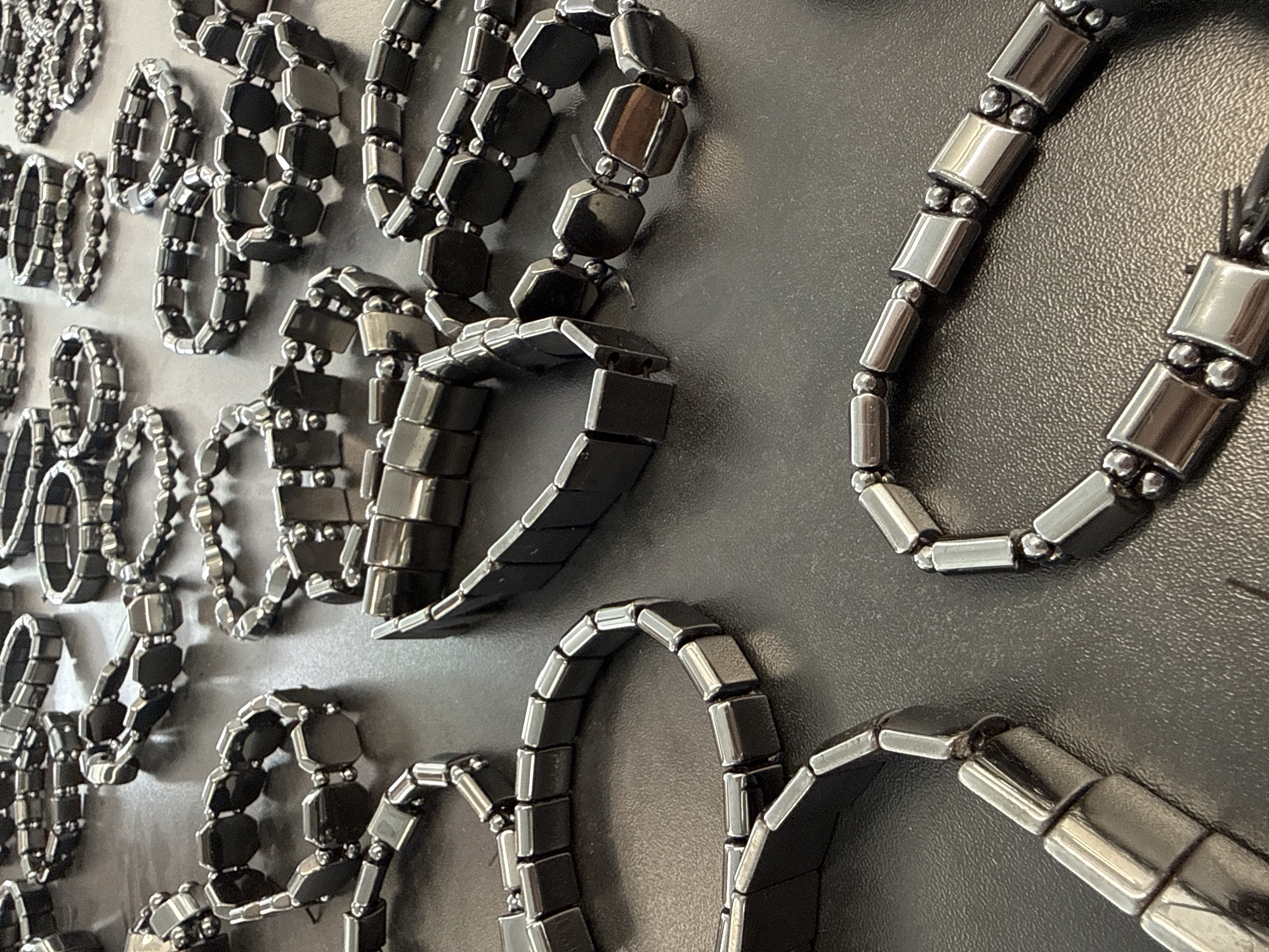 A close-up photograph shows an array of magnetic hematite bracelets displayed on a flat, dark surface. The bracelets vary in design, with some consisting of rectangular beads separated by spherical beads, and others using hexagonal or square beads. The bracelets are arranged in a seemingly random pattern, partially overlapping each other. The bracelets' surfaces reflect light, giving them a metallic sheen. The background is a plain, dark gray surface, possibly a display case or table.
I don't have information about the bracelets being in the city of Синьково, Russia.