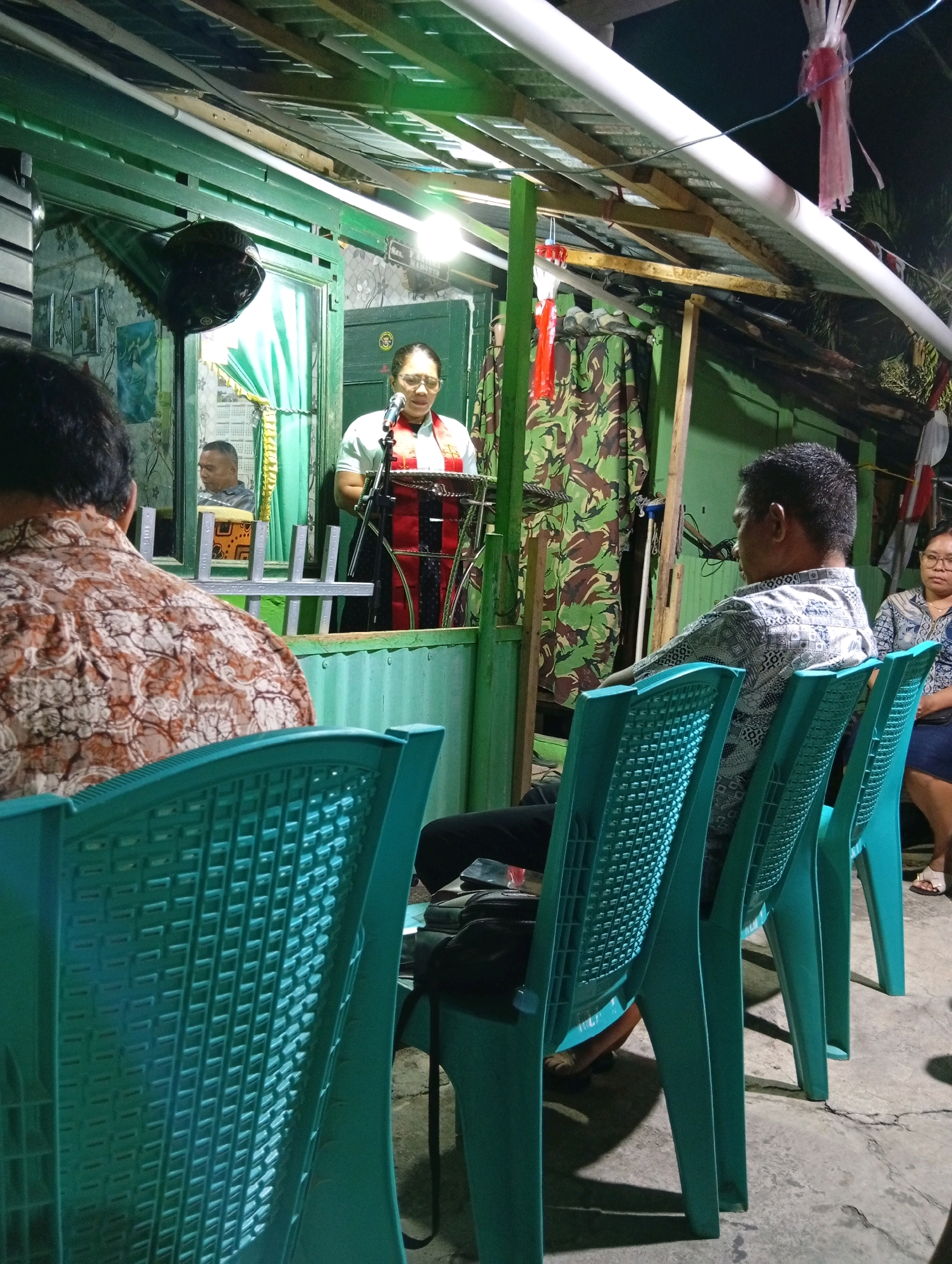 The image depicts an indoor gathering in what appears to be a community hall or a large room in Manado, Indonesia. A woman, presumably a speaker or officiant, stands at a podium with a microphone, wearing glasses and a red and black sash over a white shirt. She is facing an audience seated in light blue plastic chairs. In the foreground, the backs of several audience members are visible. To the left, a man with a patterned shirt is seated. Further back, a man is reflected in a window, also seated. The environment features green walls, a wooden ceiling structure, and camouflage-patterned fabric draped on the wall. Decorative red and white streamers are visible, suggesting a celebratory or national event. The lighting is artificial, casting shadows and illuminating the speaker.