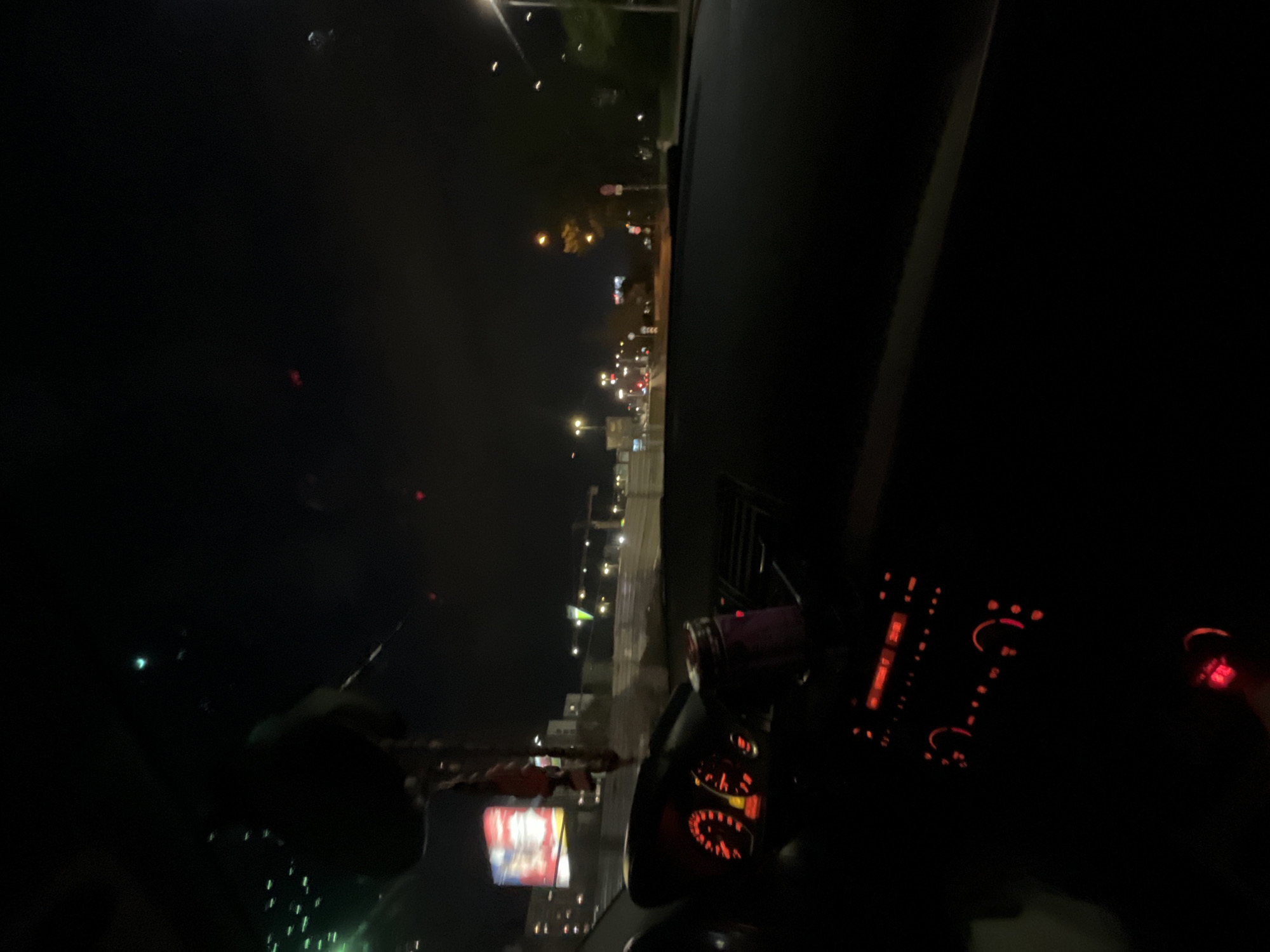 Captured from the interior of a vehicle at night, the image shows the illuminated cityscape of Sofia, Bulgaria. The dashboard and interior car elements are visible in the foreground, with red lights emanating from the instrument panel and radio. Outside, city lights stretch into the distance. The image suggests the vehicle is either stationary or moving slowly through the urban environment. A hand is visible in the bottom left of the image, along with a phone.
