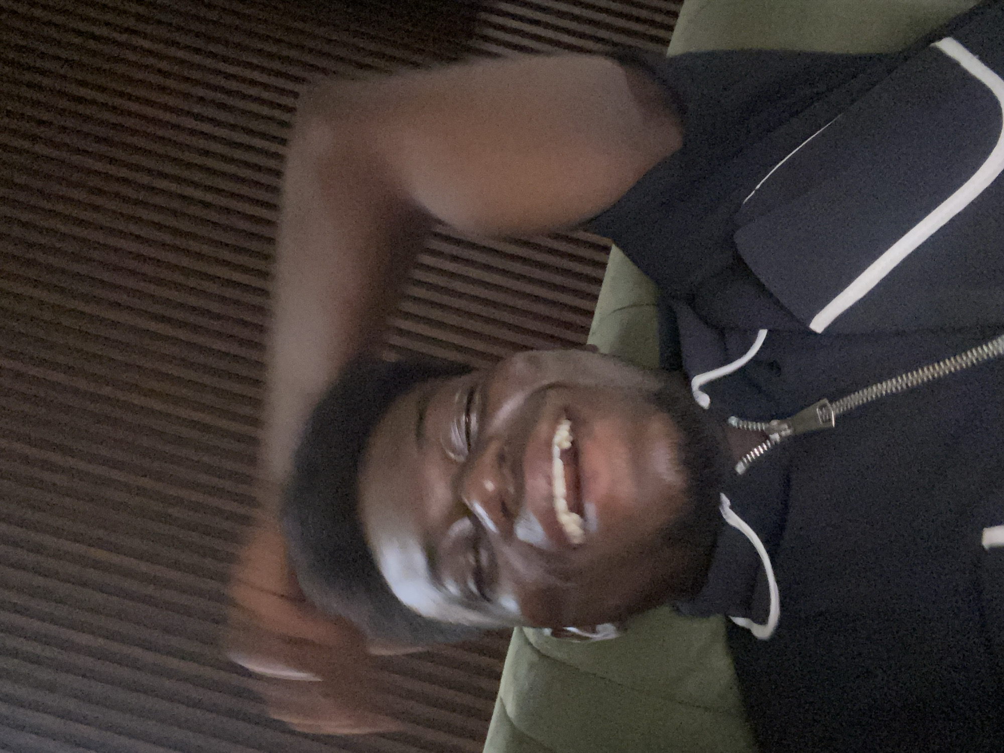 The image displays a person lying down and looking towards the camera with a joyful expression. The individual has dark skin, short dark hair, and a beard, and is captured in mid-laughter or a broad smile, with their mouth open and white teeth visible. They are wearing a dark-colored (likely black or navy blue) zippered jacket or hoodie, which features distinctive white piping or stripes along the sleeves and possibly other areas. The metallic zipper is visible down the front of the garment, partially unzipped.
The person is resting on a soft, green or olive-toned surface, which appears to be a cushion, sofa, or bed. In the background, behind the individual, there is a dark surface characterized by distinct, parallel horizontal stripes, suggesting wood paneling, a textured wall, or a ribbed fabric. The scene appears to be an indoor setting. The photograph is oriented with the person's head on the right side of the frame and their body extending upwards, indicating a 90-degree clockwise rotation of the image from a typical upright perspective. No text is visible in the image. The time of day cannot be determined from the lighting, and the specific location is not discernible beyond being indoors.
