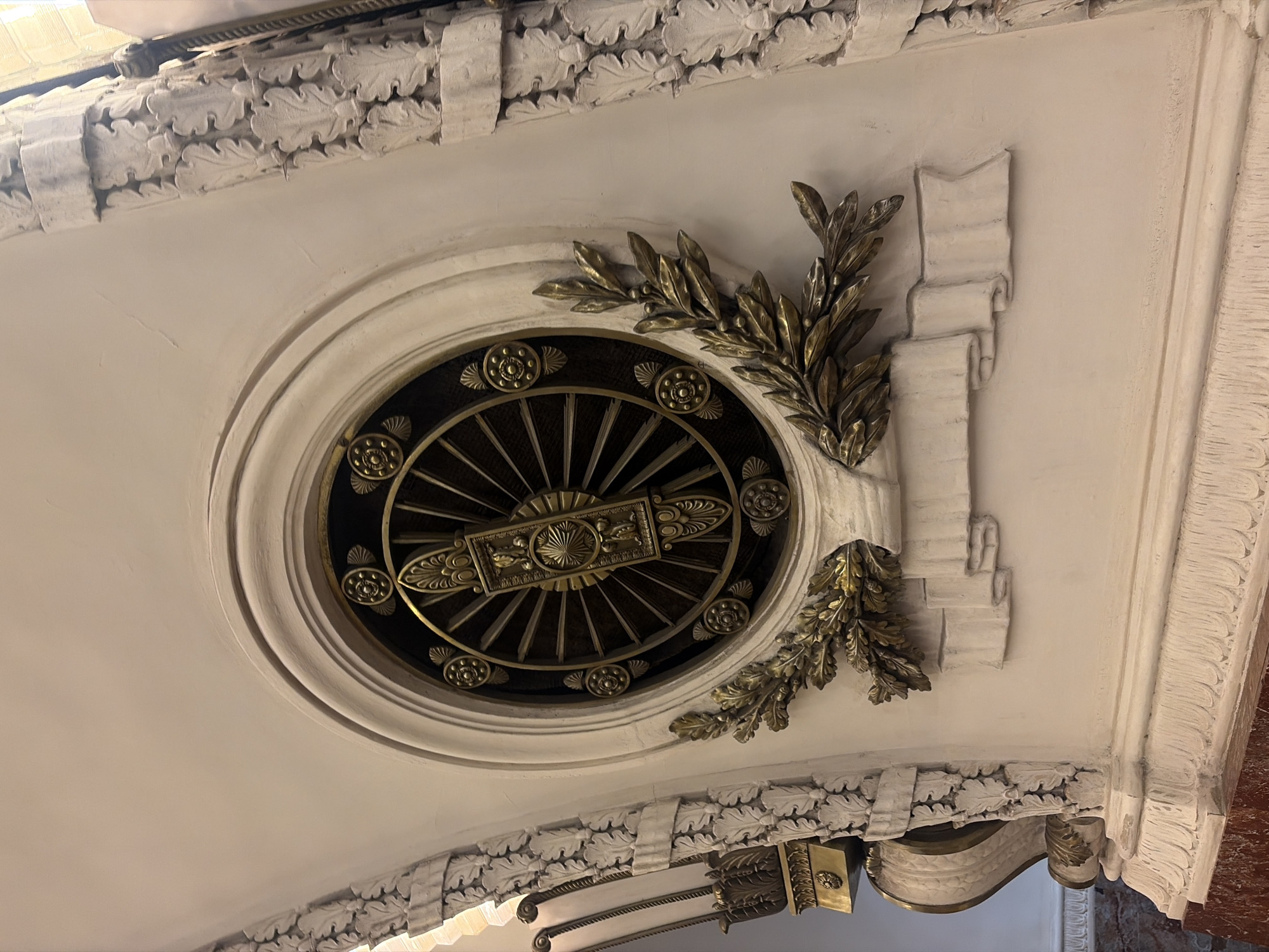 The image depicts an ornate ceiling decoration, likely found in a historic building in Saint Petersburg, Russia. The primary feature is a circular, clock-like design inset within a larger, decorative oval. The clock-like element features a series of radiating spokes emanating from a central, rectangular medallion, and is ringed by decorative rosettes. Golden metallic laurel wreaths adorn either side of the circular design. Adjacent to one of the laurel wreaths is a three tiered sculptural design that appears to mimic the rolled scroll of a document or banner. The entire oval design is set against a white ceiling, which is adorned with decorative molding in the form of stylized leaves or floral patterns. The image is taken from a low angle, looking upwards towards the ceiling. Portions of a staircase and a marble column can be seen in the background. The overall style is reminiscent of classical architectural ornamentation.
