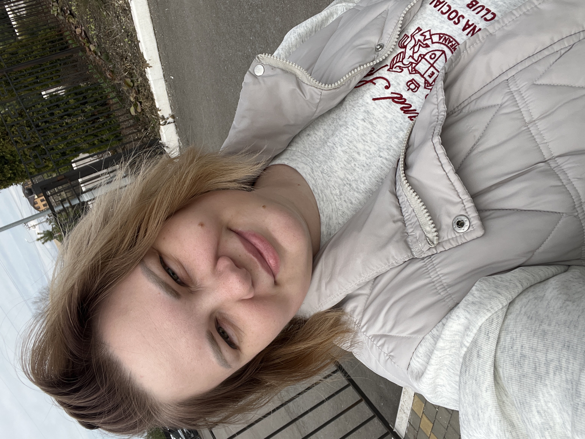 A woman is taking a selfie outdoors in Usman, Russia. The background consists of a sidewalk with yellow and grey pavers, a road, a low black metal fence, and some greenery. The woman is wearing a light grey quilted vest over a grey sweatshirt that has a red and white emblem on it. She has light brown hair.