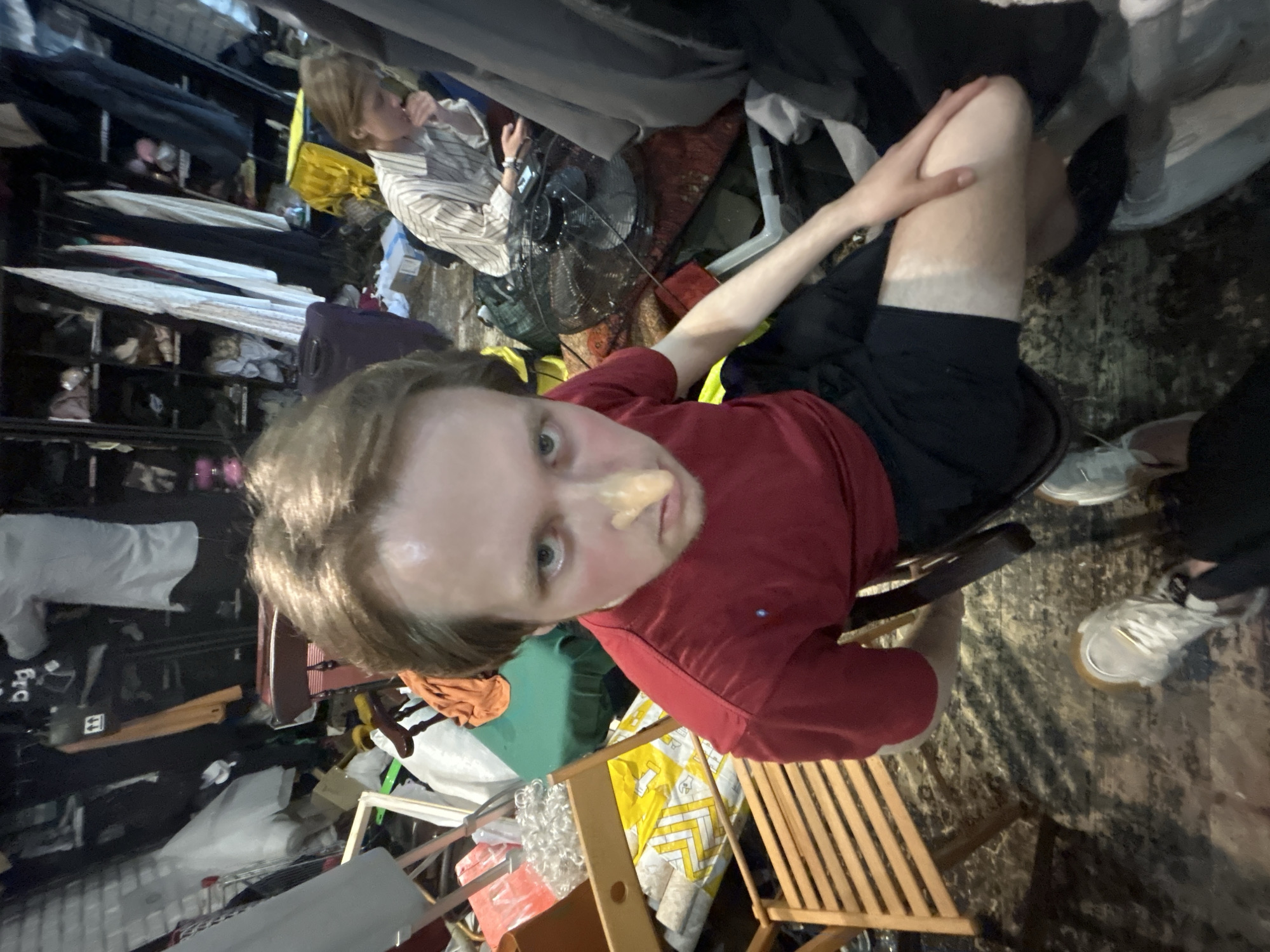 A high-angle shot depicts a person with light skin, wearing a red shirt and black shorts, seated on a wooden chair. A substance is visible on their nose. A tan line is visible on their leg.
In the background is a person in a striped shirt using an electronic device. A floor fan is situated nearby. Clothing racks and assorted items contribute to a cluttered environment. The exact location is not identifiable from the given image.