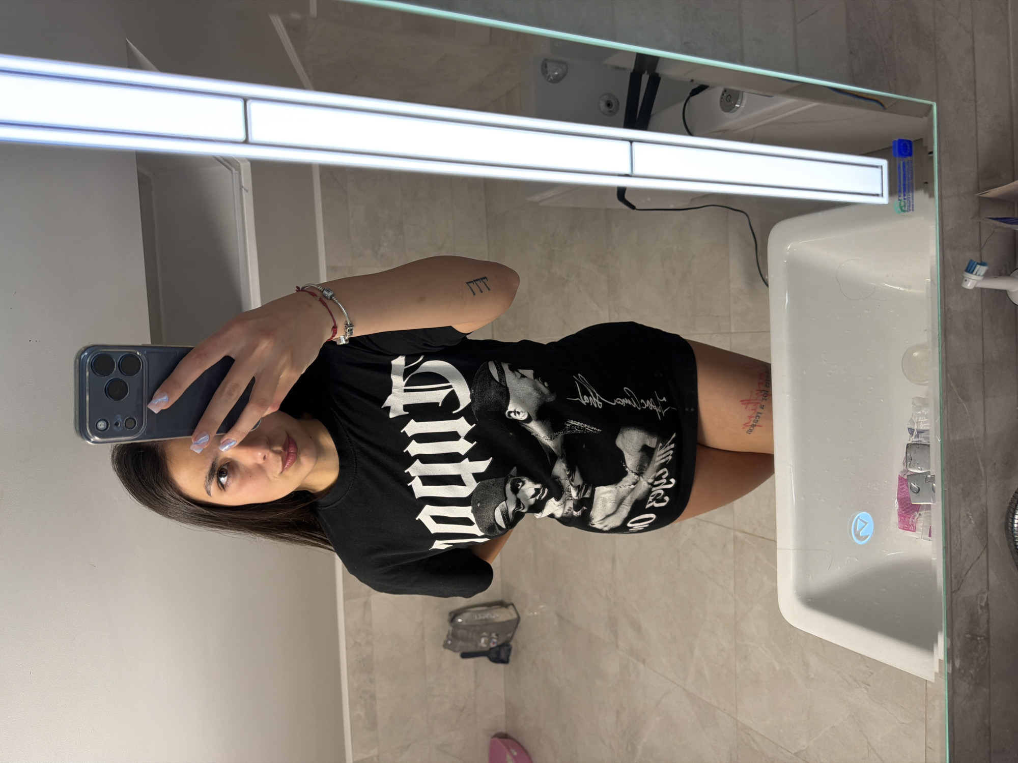 A selfie is taken in a bathroom with light gray tiled walls and flooring. A woman is holding a smartphone in her left hand to take the picture, her arm visible in the frame. She is wearing a black t-shirt with white print and short sleeves. The background includes a sink, a mirror with a built-in light fixture, and a toilet. The exact location in the city of Gabrovo, Bulgaria, cannot be determined from the given image.