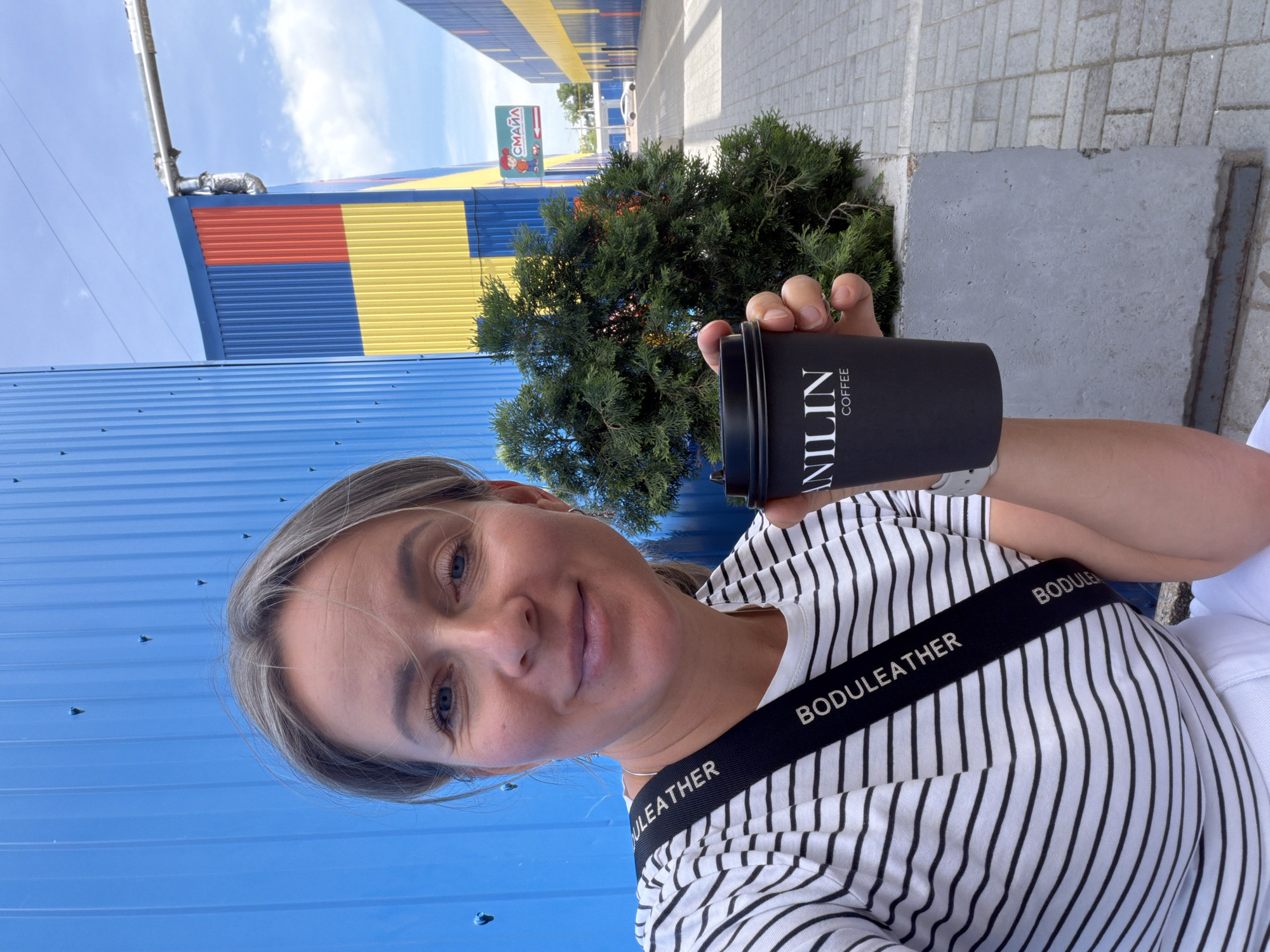 The image shows a selfie of a woman holding a black VANILIN Coffee cup. She is wearing a striped shirt with a strap across her shoulder that says "BODULEATHER." Behind her is a small evergreen bush and sections of a building that are blue, yellow, and red. A sign is visible further down the street that says "СМАЙА" (SMAIA) in Cyrillic. The scene appears to be set in an urban environment. Based on the sign, it is in the city of Chaikovsky, Russia.