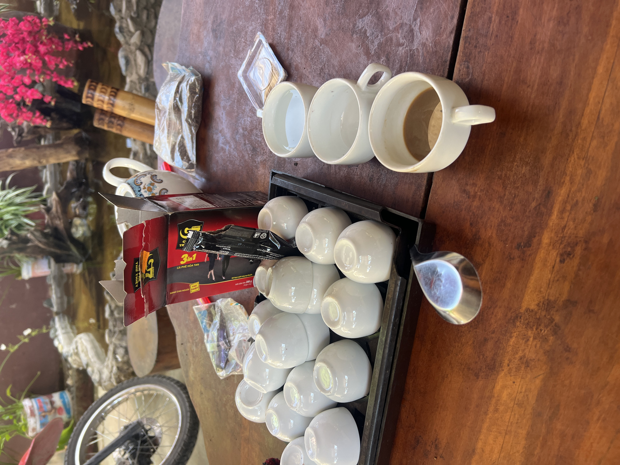 In Lùng Tám Commune, Vietnam, a wooden table is set for a tea or coffee ceremony. A tray on the table holds numerous small, white ceramic cups, some stacked and others nested. To the right, three larger white ceramic mugs are present; two contain clear liquid, likely water, and the third holds a dark brown beverage, possibly coffee. Beside the mugs, a small, clear plastic container rests on the table. A red and black cardboard box, labeled with Vietnamese text and graphics that appear to be for coffee, is open and placed prominently on the table, with a black packet emerging from it. Next to the box, a white ceramic teapot with a floral pattern and a handle sits near two bamboo cylinders. In the background, a small pond with decorative rocks and plants is visible, along with a portion of a motorcycle wheel and tire on the left side of the image. The overall scene suggests a moment of shared refreshment in an outdoor setting.