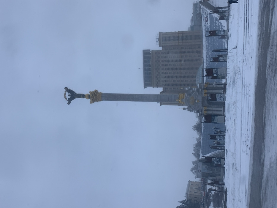 The image displays Maidan Nezalezhnosti (Independence Square) in Kyiv, Ukraine, under snowy, overcast winter conditions. Centrally positioned is the Monument to Independence of Ukraine, a tall grey column with a gilded capital, topped by a dark-colored statue of a winged female figure (Berehynia) holding a wreath. Behind the monument, the multi-story Hotel Ukraine building is visible, featuring a "HOTEL" sign on its upper right facade. Below the monument and extending across the mid-ground are the snow-covered structures of the Globus Shopping Mall entrance, characterized by glass panels and banners. The entire ground level, including steps and walkways, is blanketed in snow. The sky is a uniform, light grey, indicating heavy cloud cover or active snowfall, with individual snowflakes discernible. One person, dressed in dark clothing, walks on the snow-covered ground on the far right side of the frame, moving away from the center.