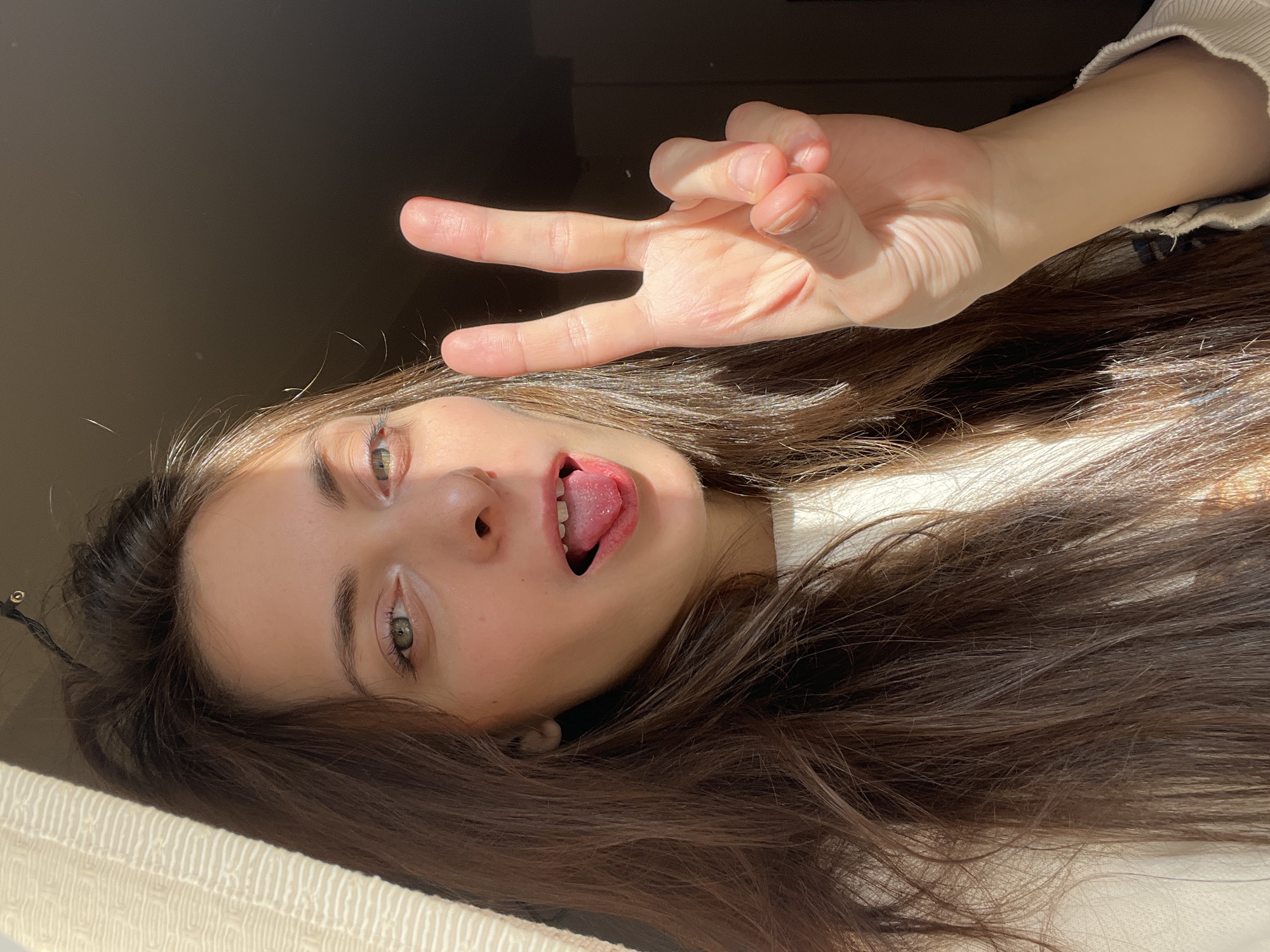 A young woman lies on her back with her head near a window. Her brown hair is spread out around her. She is making a peace sign with her right hand, and her tongue is sticking out. The background is blurred, but appears to be a room with natural light. There is no evidence to suggest the location is in Krasnoyarsk, Russia.