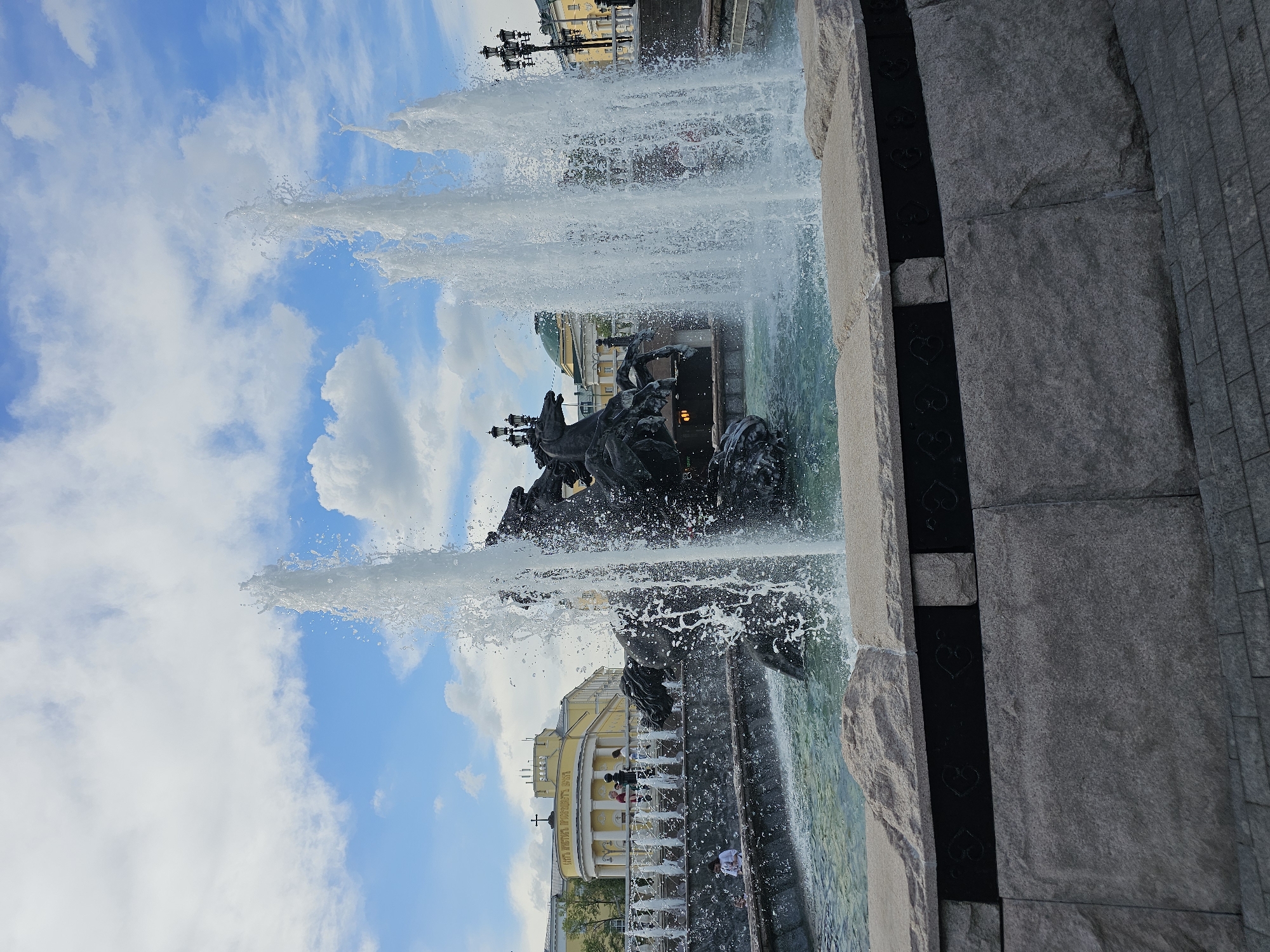 The image captures the Friendship of Nations Fountain, located in Moscow, Russia. The fountain features dramatic water jets shooting high into the air, set against a backdrop of a blue sky with scattered clouds. The fountain's sculptural elements, dark in color, depict various figures and symbols. The structure of the fountain is made of concrete and stone, with some decorative black metalwork featuring heart-shaped motifs. In the background, a light-yellow building with white columns and a green dome can be seen. There are some people walking around the edges of the fountain. The image is oriented vertically.