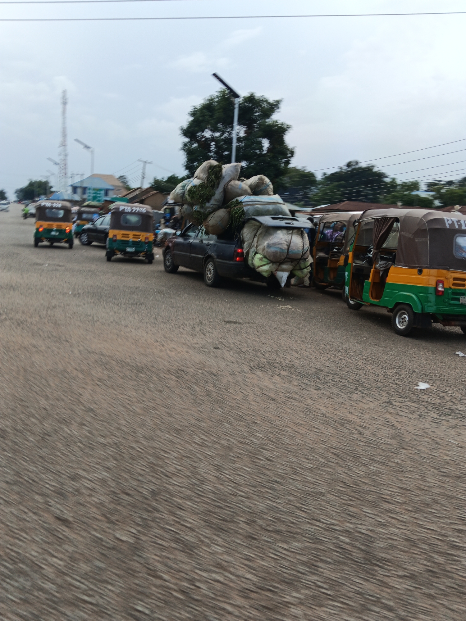 In Bukuru, Nigeria, a traffic scene unfolds on a paved road under an overcast sky. The foreground is dominated by the textured surface of the road, with a slight motion blur suggesting movement.
Several vehicles are present, creating a sense of congestion. In the middle ground, a dark-colored car is heavily laden with large, round bundles wrapped in what appears to be burlap or fabric, piled high on its roof and trunk. These bundles are secured with ropes and netting. Following the car are at least three auto-rickshaws, also known as "keke napeps." The closest one is a yellow and green vehicle with a brown canopy. The other two are visible behind it, one yellow and green, and another mostly obscured. The auto-rickshaws also appear to be carrying goods or passengers, though the details are less clear.
In the background, buildings and trees line the sides of the road. A tall communication tower is visible on the left, with streetlights and overhead power lines crisscrossing the sky. The architecture of the buildings suggests a commercial or residential area. The overall impression is one of everyday life and commerce on a Nigerian roadway.