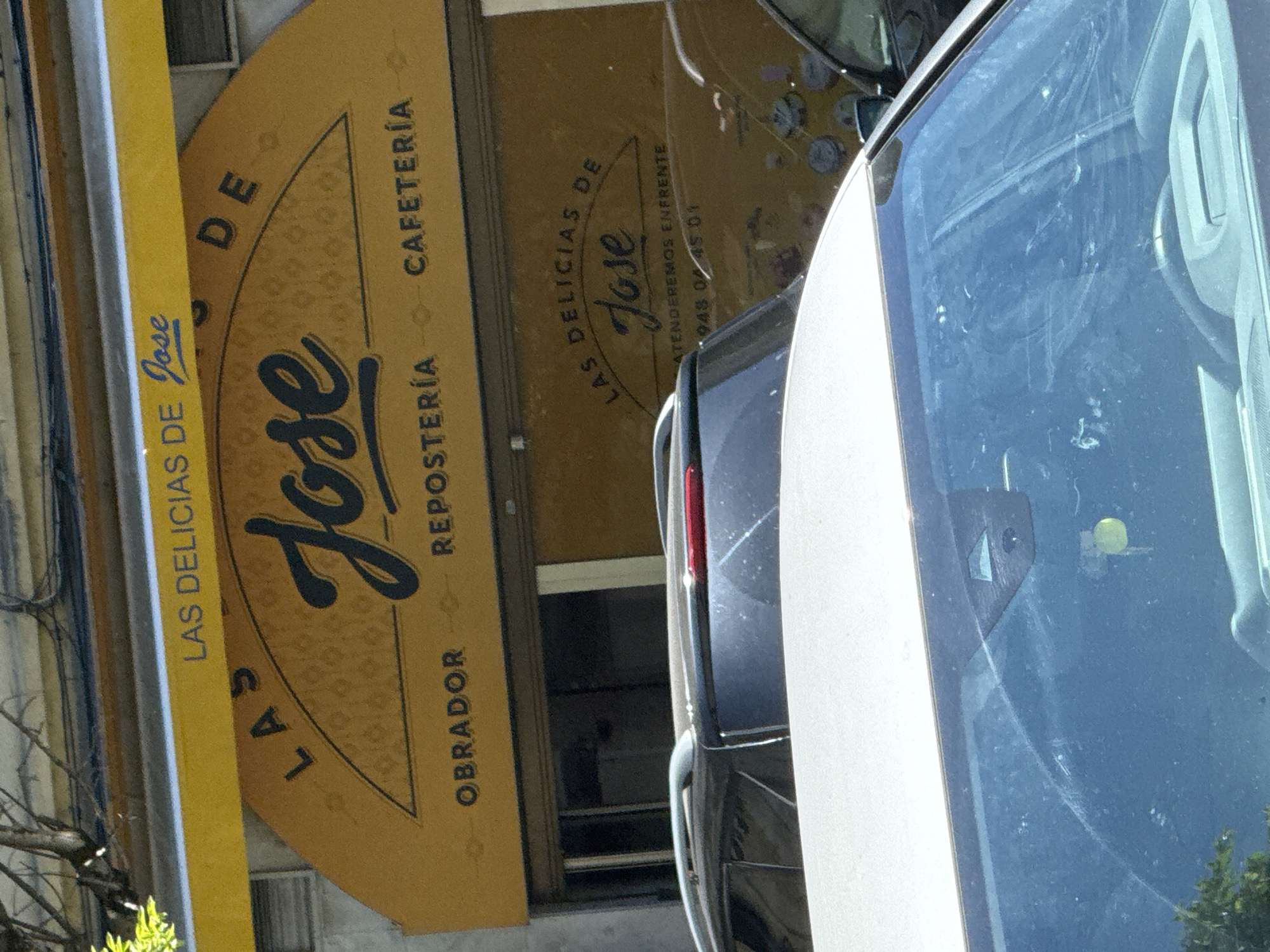 The image shows a storefront for "LAS DELICIAS DE JOSE," a business that appears to be a bakery and cafe. The storefront features a large yellow awning with the business name in blue lettering. Below the awning, there is a curved sign with the same name, "LAS DELICIAS DE JOSE," in dark lettering. The words "OBRADOR," "REPOSTERÍA," and "CAFETERÍA" are also visible on the sign, indicating the services offered. The reflection on the storefront glass reveals a portion of the interior, including closed shutters and possibly displays. In the foreground, the hoods and windshields of two cars are visible, suggesting the photo was taken from a street or parking area. The overall impression is an urban commercial setting, likely in a city. Without further context, the specific location in Pamplona, Spain, cannot be definitively determined. However, based on the signage, it is a commercial establishment focused on pastries and coffee.