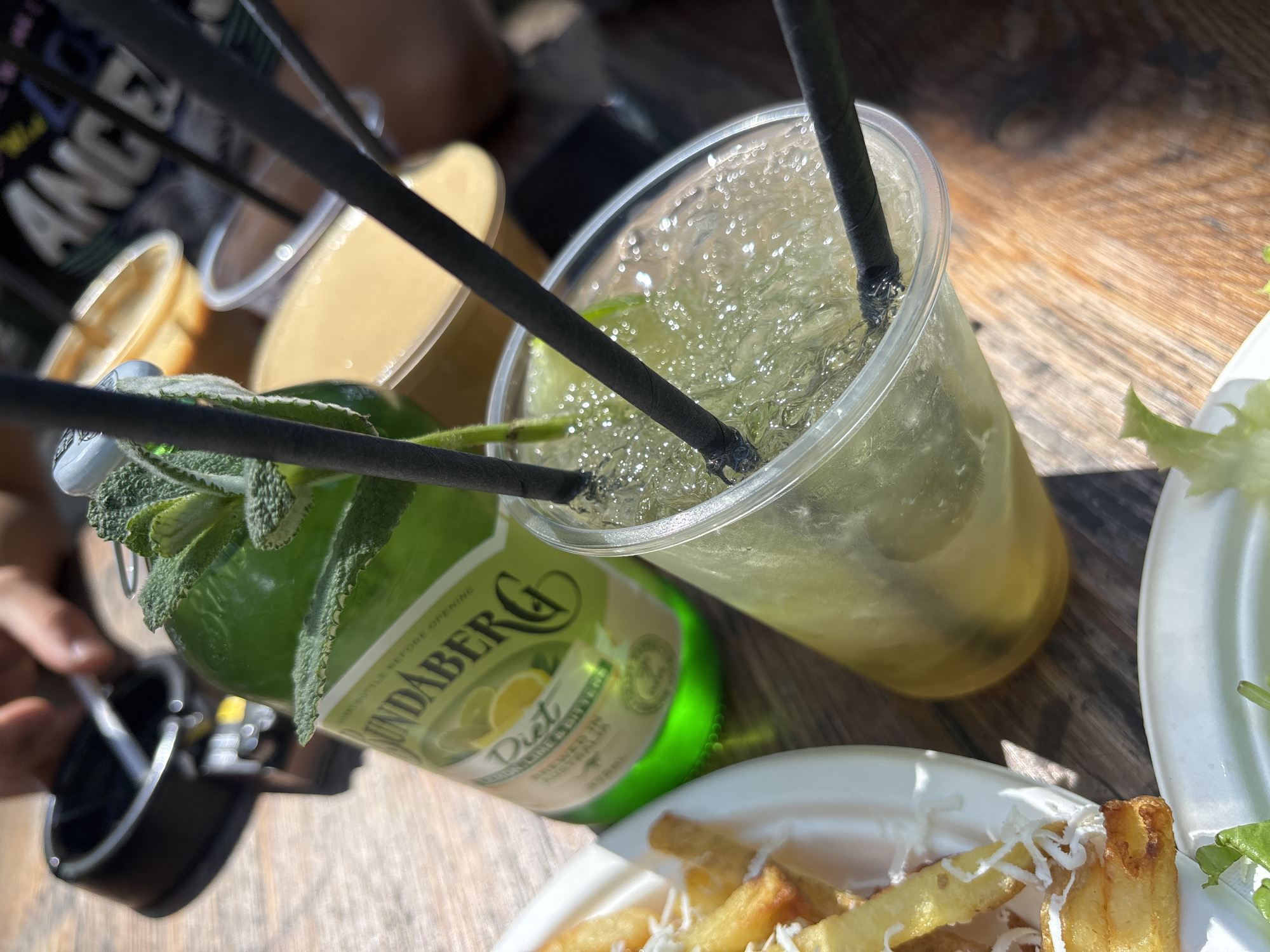 The image shows a close-up of a table setting outdoors. The table appears to be made of wood.
Items on the table include:
* A plastic cup filled with a clear, bubbly drink and ice, two black straws are inserted.
* A green glass bottle labeled "Bundaberg Diet Lemon Lime & Bitters". The bottle has a sage plant growing on it.
* A white plate of French fries with grated cheese.
* A white plate of salad.
* A few plastic cups with a yellow liquid.
* A black camera and other personal belongings are visible near the left edge of the image.
The location is likely outdoors at a restaurant or cafe due to the presence of drinks and food. The image lacks geographical indicators for Sofia, Bulgaria.