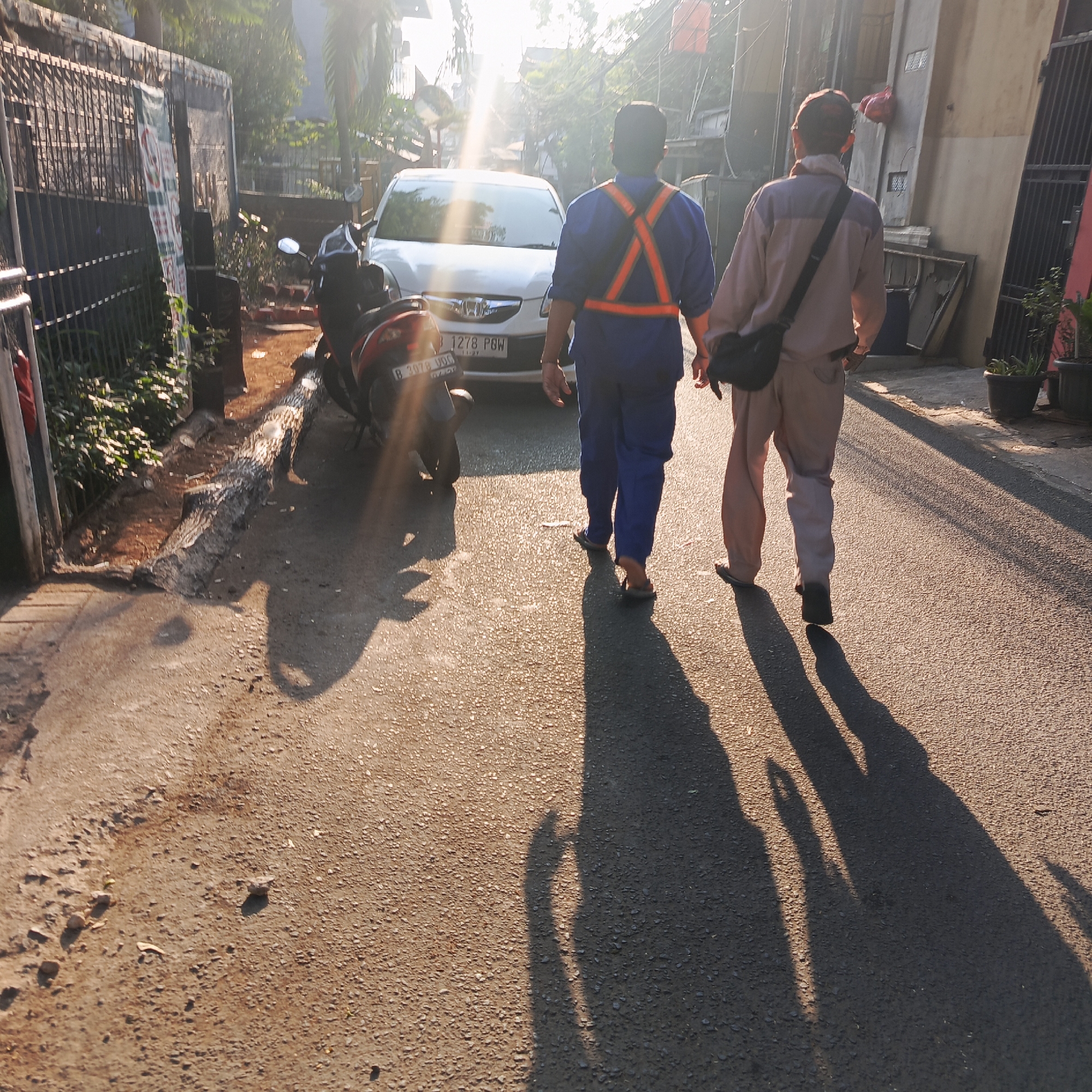 Two men walk away from the camera down a paved street in South Jakarta, Indonesia. The man on the left wears a blue uniform with an orange reflective vest. The man on the right wears a light-colored long-sleeved shirt and matching pants, with a black messenger bag slung across his chest. A white car is parked on the street to their left, and a motorcycle is parked to the left of the car. The sun is low in the sky, casting long shadows of the men and the car onto the street. The environment appears to be a residential area with buildings and vegetation visible in the background. The street is lined with fences and potted plants.