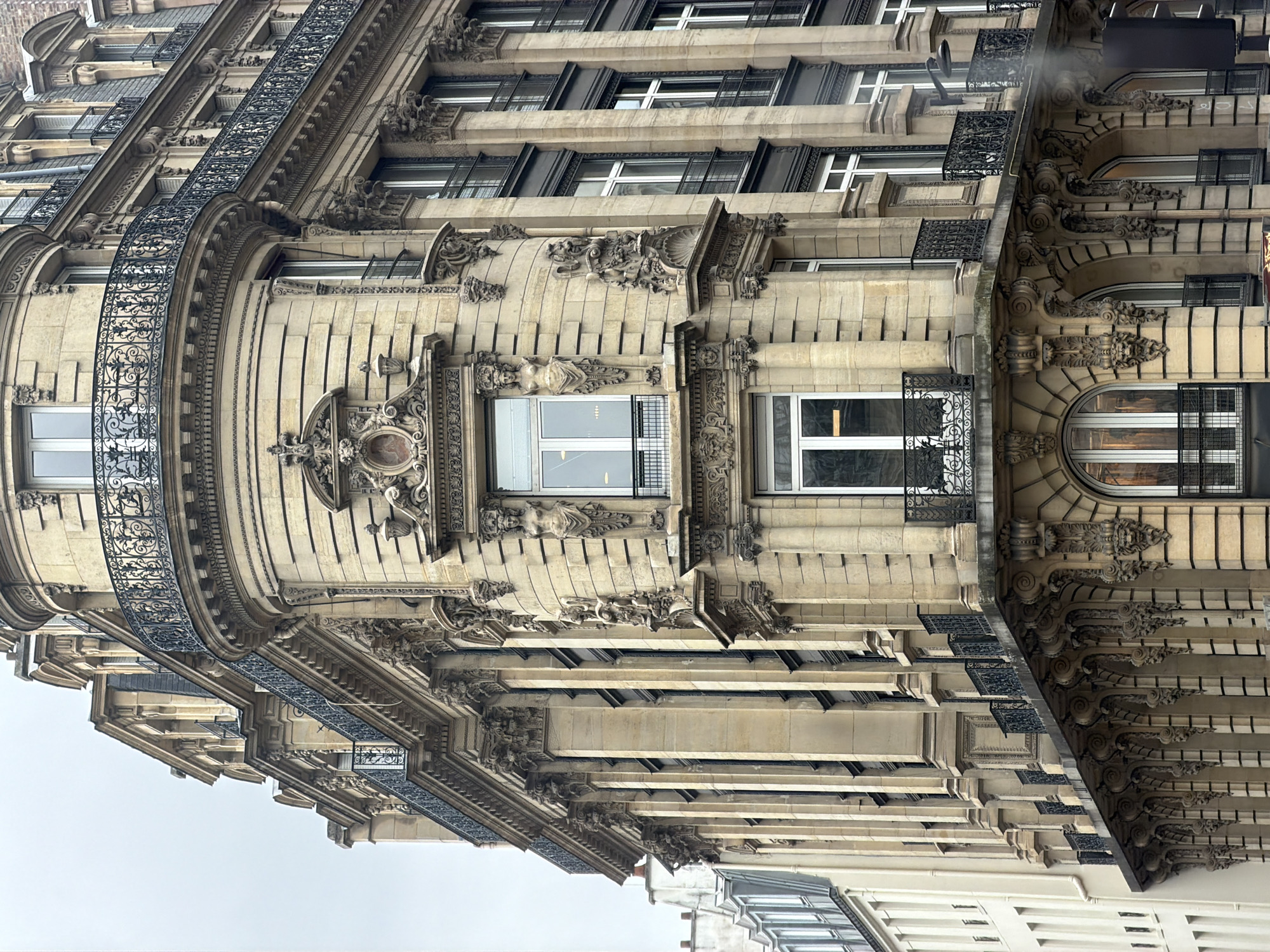 The image depicts the exterior of a Parisian building. It features detailed architectural elements such as ornate moldings, decorative facades, and wrought-iron balconies. The building's architecture suggests it is from the late 19th or early 20th century, with a light-colored stone facade and varied window styles. The angle suggests a street view looking upwards at the structure. The sky is overcast. There are no people visible. The building is located in Paris, France, based on its architectural style.