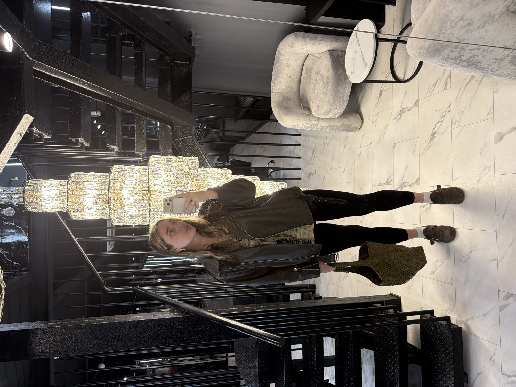 A young woman takes a full-length selfie in an upscale interior space, possibly a hotel lobby or a showroom in Moscow, Russia. She is positioned next to a large, industrial-style black metal staircase with diamond plate steps and sturdy railings, which dominates the left side of the frame.
The woman wears a dark brown leather jacket over a light-colored, textured knit top, black leggings, dark brown flat shoes with buckle details, and white ankle socks. In her left hand, she holds a green, intricately woven tote bag. She holds a light-colored smartphone with multiple cameras up to capture her reflection, gazing slightly upwards with a neutral expression.
To her left, a magnificent multi-tiered chandelier, composed of numerous small crystal or glass elements, hangs prominently, emitting a warm, bright light. This light creates reflections on her phone screen and a black marble-patterned wall panel near the staircase. The floor is covered with large, rectangular white tiles featuring grey marble-like veining. In the background, to the right, light grey upholstered armchairs with unique, organic shapes are visible alongside a round table with a white marble top and a black metal base.
The scene suggests a modern and luxurious environment, blending raw industrial elements from the staircase with opulent and contemporary furnishings. There is no visible text in the image.