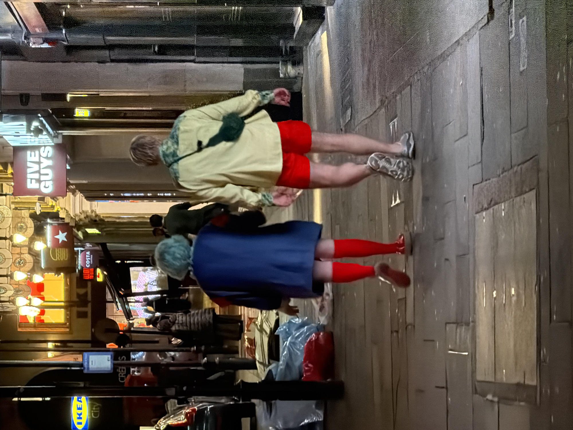 A candid, street-level, medium shot depicts two individuals walking on a paved pedestrian walkway in what appears to be a back alley in London. The individual in the foreground is wearing a blue dress, red tights, and red shoes. The individual in the background is wearing a light yellow jacket, red shorts, and sneakers. They are walking in the same direction, away from the viewer. In the background, a variety of retail signs are visible, including "FIVE GUYS," "COSTA COFFEE," and "IKEA City." The street is lined with retail stores and overhead lighting.