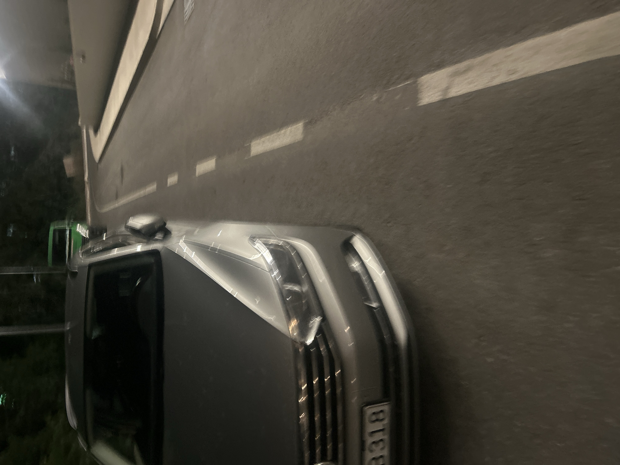 In Boreti, Montenegro, at night, a silver car is parked on a road with white dashed lines. The car's front is visible, showing its grille, headlights, and license plate which reads "3318". In the background, a green bus is also parked on the road. The environment appears to be a street with trees and a concrete structure on the right side. The lighting suggests artificial illumination from streetlights or nearby buildings. The scene is static, with no visible activity or interaction.