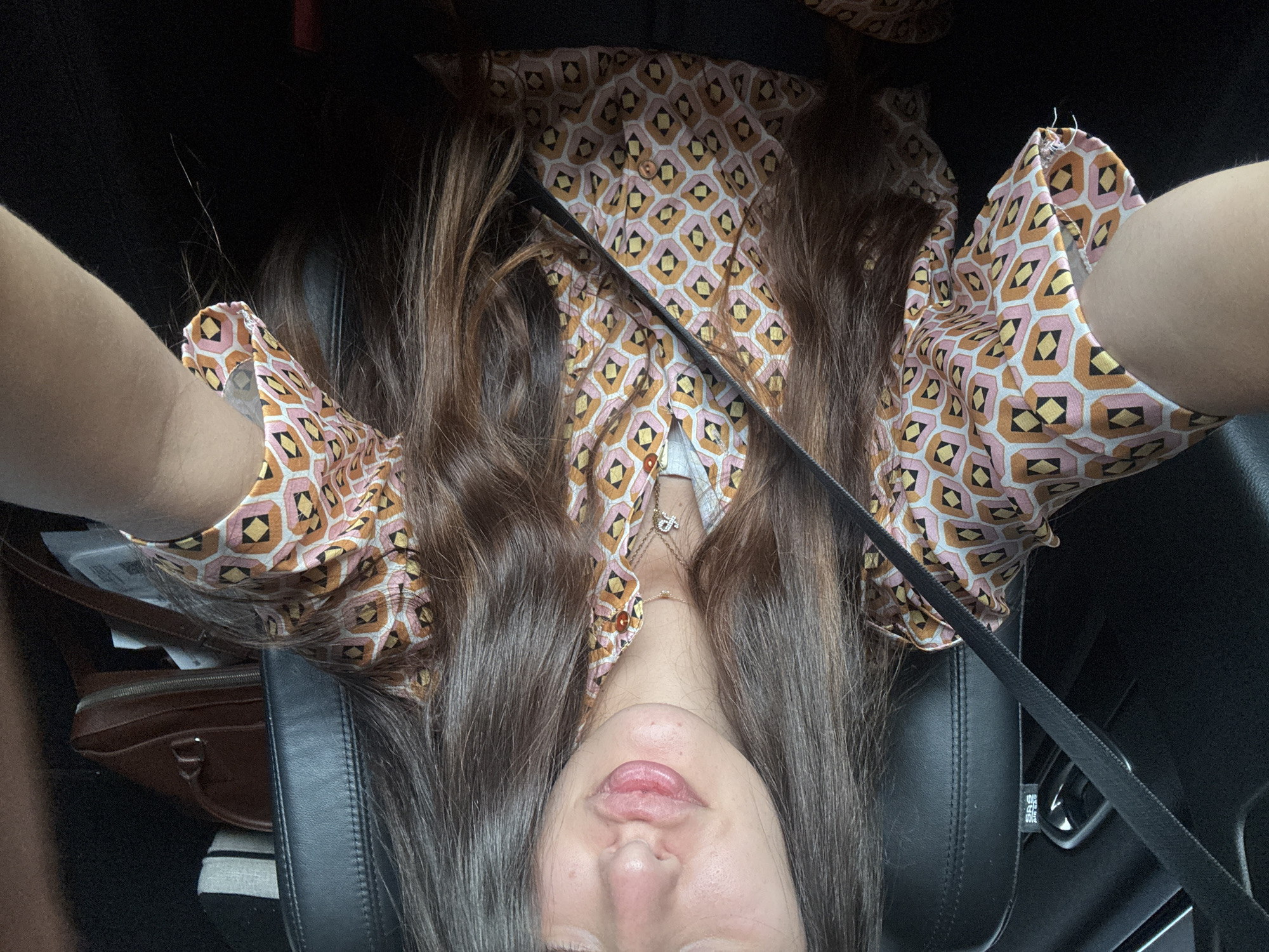 A young woman with long, dark brown hair is sitting in the driver's seat of a car, wearing a pink, orange, and yellow patterned button-down shirt. She is looking towards the camera with her lips pursed. A black seatbelt is visible across her chest. The car's interior is dark, with black leather seats and a glimpse of a brown purse on the passenger seat. The image was taken in Deerfield Beach, United States of America.