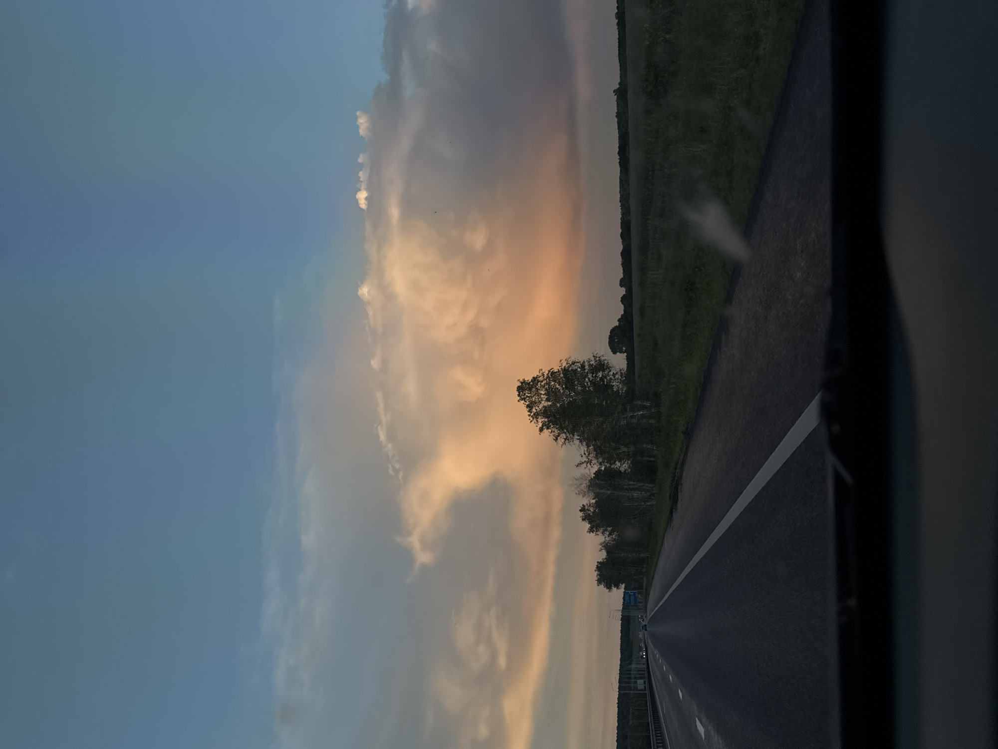 The image depicts a view from inside a vehicle, likely on a highway, in Voskresensky District, Russia. The road stretches into the distance with a single white line marking lanes. A field borders the road on one side, and trees are visible in the mid-ground. The sky is mostly blue with large, illuminated clouds that appear to be sunset or sunrise colors, probably from sunlight. A portion of the vehicle's interior is seen in the bottom-right corner.