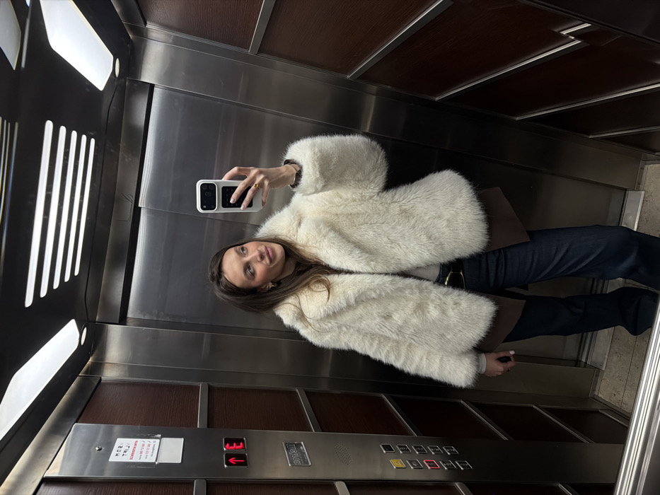 An elevator interior in Budva, Montenegro, is depicted. The environment consists of brushed stainless steel walls, dark wood paneling, and a control panel on the left. The control panel features illuminated red up-arrow and 'E' buttons, along with additional unlit numerical and functional buttons. A partially visible sign displaying Cyrillic script, including "МОЖ ТРИ!", and the letters "MONTE" are present on the control panel, consistent with the specified location. The ceiling incorporates recessed lighting fixtures.
A single female subject is shown taking a mirror selfie. She has long, dark brown hair and is wearing a cream-colored faux fur coat over a dark brown blazer, a light-colored top, a dark belt with a gold buckle, and dark blue trousers. Her right hand, adorned with white painted fingernails and a gold-colored ring, holds a white smartphone with a black camera module, positioned for the photograph. The subject is looking slightly to her right, exhibiting a subtle smile. The floor is a light-colored hard surface.