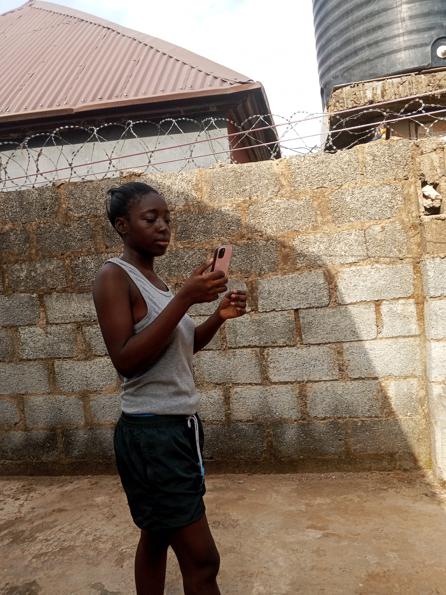 A young woman stands outdoors, holding a pink cell phone in her hands. She wears a gray tank top and dark green shorts with a light-colored drawstring. The background consists of a gray brick wall topped with barbed wire. A brown roof is visible above the wall on the left, and a black water tank is seen on the right. The ground appears to be a concrete surface.
The image was taken in Abuja, Nigeria.