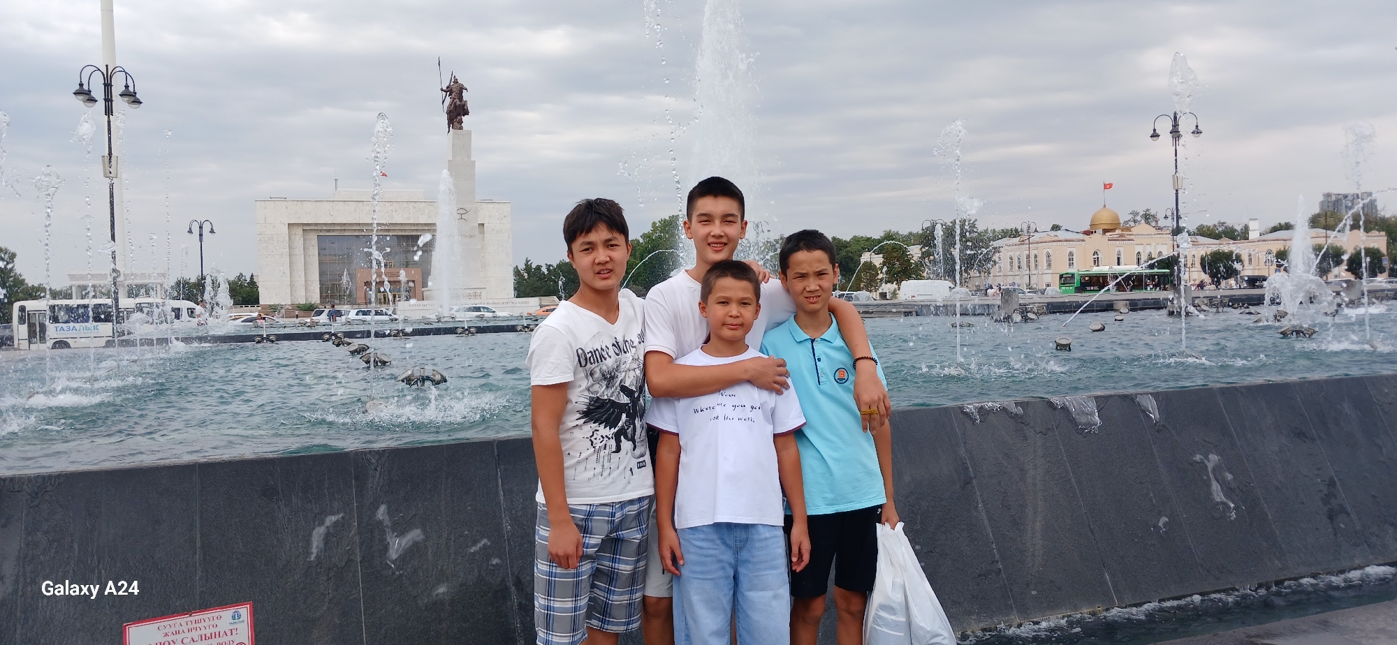 The image shows four boys posing in front of a large, active fountain in Bishkek City, Kyrgyzstan. The fountain is in the foreground with water spraying upwards. In the background, behind the fountain, is a monumental statue of a rider on a horse, a white building with a columned entrance, and a smaller building with a golden dome and a flag on top. Streetlights and trees are also visible. The sky is overcast. The boys are of various ages and are casually dressed. One boy on the far left wears a white t-shirt with a graphic print and plaid shorts. The boy next to him, in the center, wears a white t-shirt with writing and denim shorts. To his right, another boy wears a light blue polo shirt and black shorts. The boy on the far right, partially obscured, wears a white t-shirt. The overall scene appears to be a public space in the city.