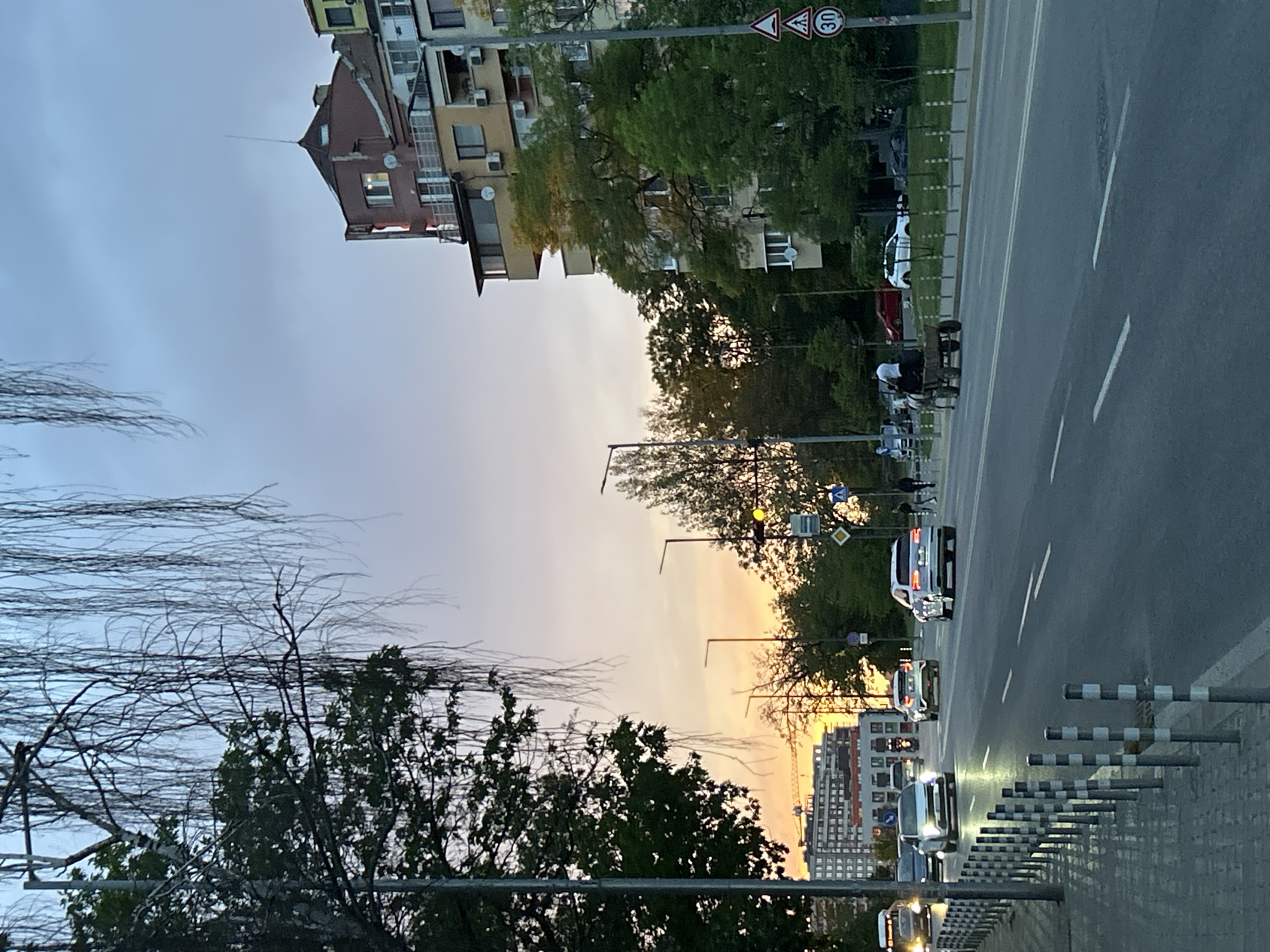 The image depicts a street scene in Sofia, Bulgaria at dusk. The sky transitions from pale blue to orange near the horizon. Tall buildings and trees line both sides of the road. A multi-lane road with white lines and striped poles on the side is visible, with several cars driving in both directions. A street light is illuminated with a yellow light. Pedestrians are walking along the sidewalks. Street signs are mounted near trees.