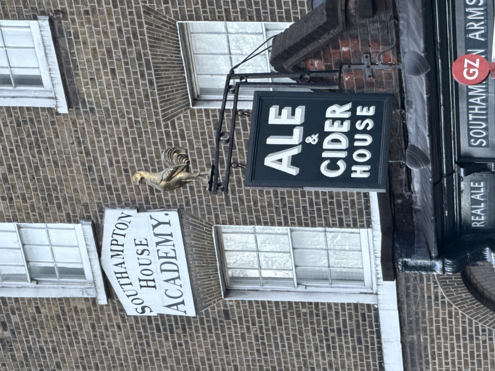 The image depicts a building in London, United Kingdom, featuring a brick facade. Signage on the building identifies it as "Southampton House Academy" and the "Southam GZN Arms," indicating it is both an educational institution and a pub. Additional signage advertises "Ale & Cider House" and "Real Ale." A decorative golden bird statue is positioned near the "Ale & Cider House" sign. The scene includes windows with white frames, and the image appears to be taken during daylight hours.