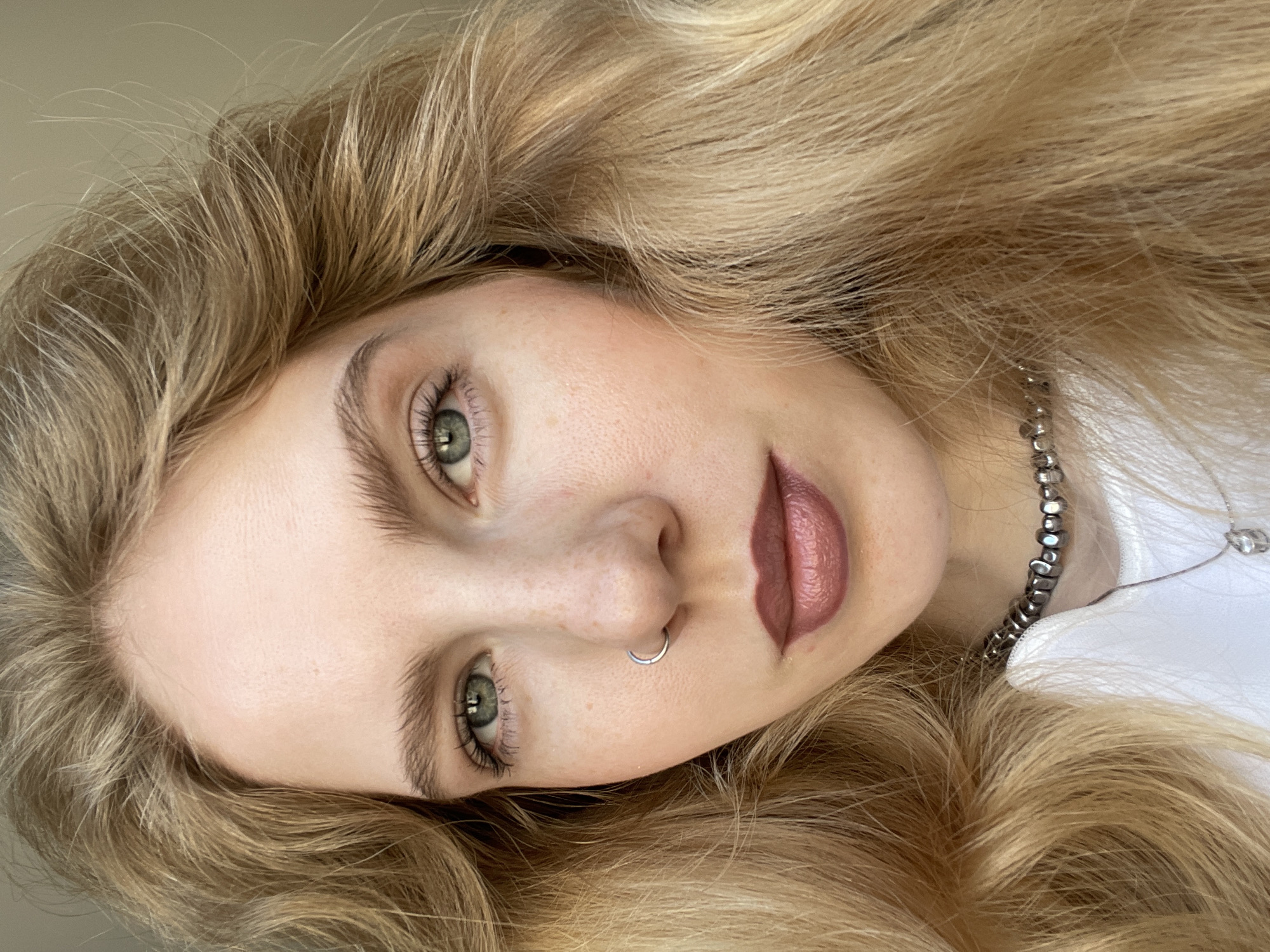 A close-up, angled portrait shows a fair-skinned woman with blonde, wavy hair. Her eyes are green, and she has a silver nose ring. She is wearing a muted mauve-colored lipstick. A silver beaded necklace is visible on her neck, against a white shirt. The background appears to be a neutral wall. There is no indication of the location in Izhevsk, Russia, or any other city. The focus is entirely on the woman's face and upper neck.