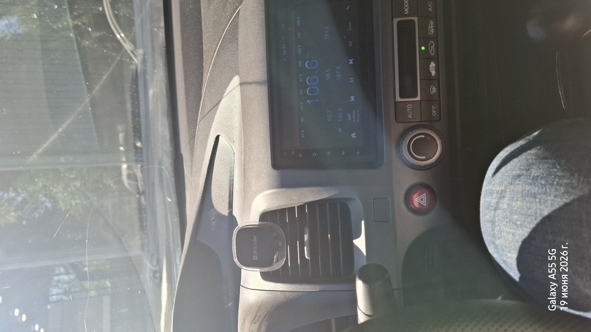 This media file is a photograph taken from inside a car, looking towards the dashboard. The sun is shining, creating bright highlights and shadows on the dashboard and windshield, suggesting it is daytime. The car's interior appears to be in good condition, with a modern touchscreen infotainment system displaying radio frequencies and controls. A mobile phone mount with the brand "defender" is attached to an air vent. To the right of the infotainment system, climate control buttons and a hazard light button are visible. The lower right of the image shows a portion of a person's denim-clad leg, likely the driver or a passenger. A timestamp and device information in Russian indicate the photo was taken on June 19, 2026, at 16:37. The location is identified as Rostov-on-Don, Russia. The overall scene depicts a routine moment inside a parked or driving car on a sunny day.