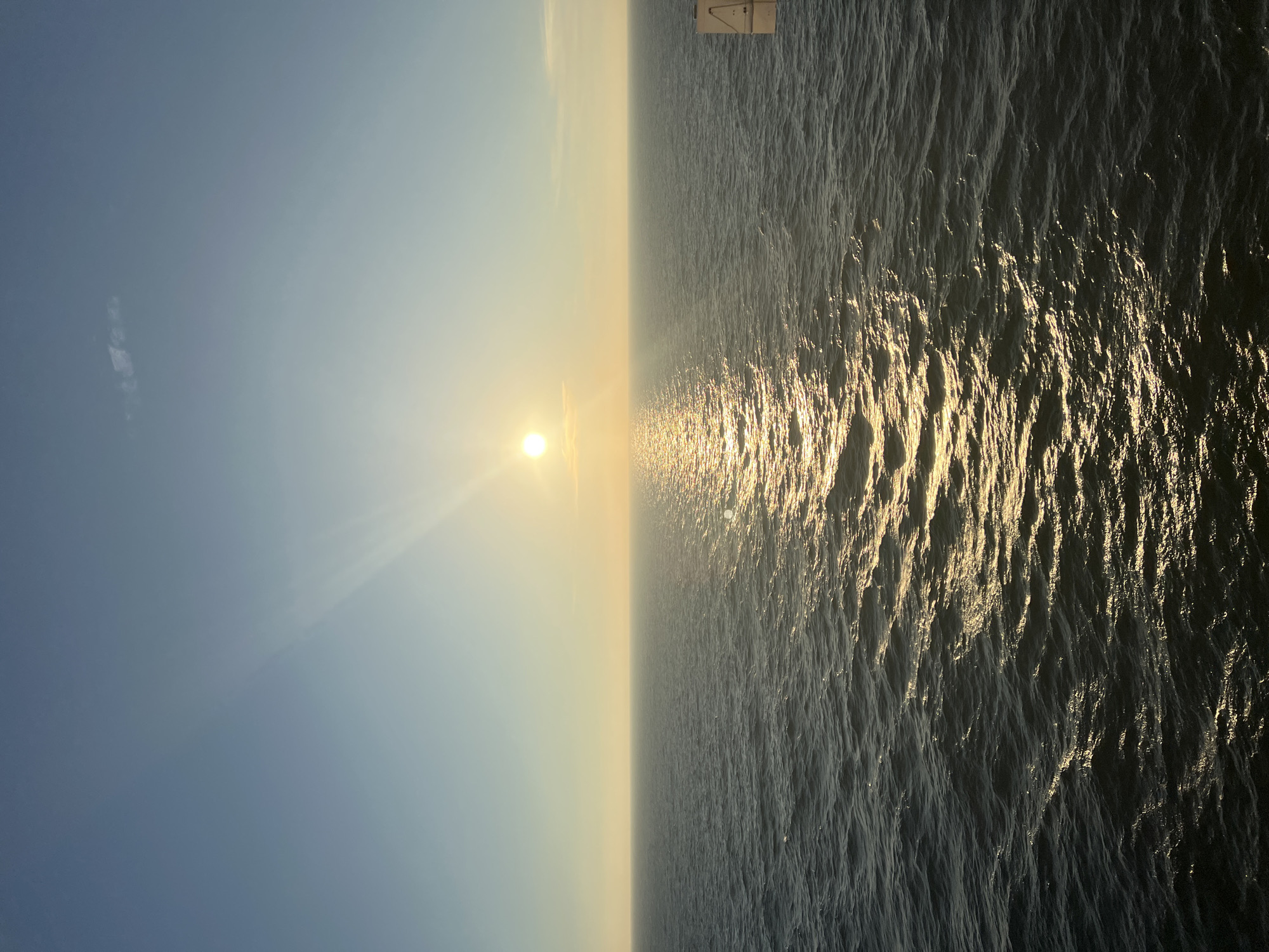 A serene ocean view at sunset is depicted, with the sun low on the horizon casting a golden path across the rippling water. The sky transitions from a clear, pale blue at the top to a soft yellow near the horizon, with a few wispy clouds. The ocean is a deep, dark blue, with small waves creating a textured surface. The overall scene conveys a sense of calm and vastness. There are no people or discernible structures in the immediate foreground or mid-ground, though a small, white sign with some markings is visible on the far right edge of the image, suggesting the presence of a vessel or offshore platform.
