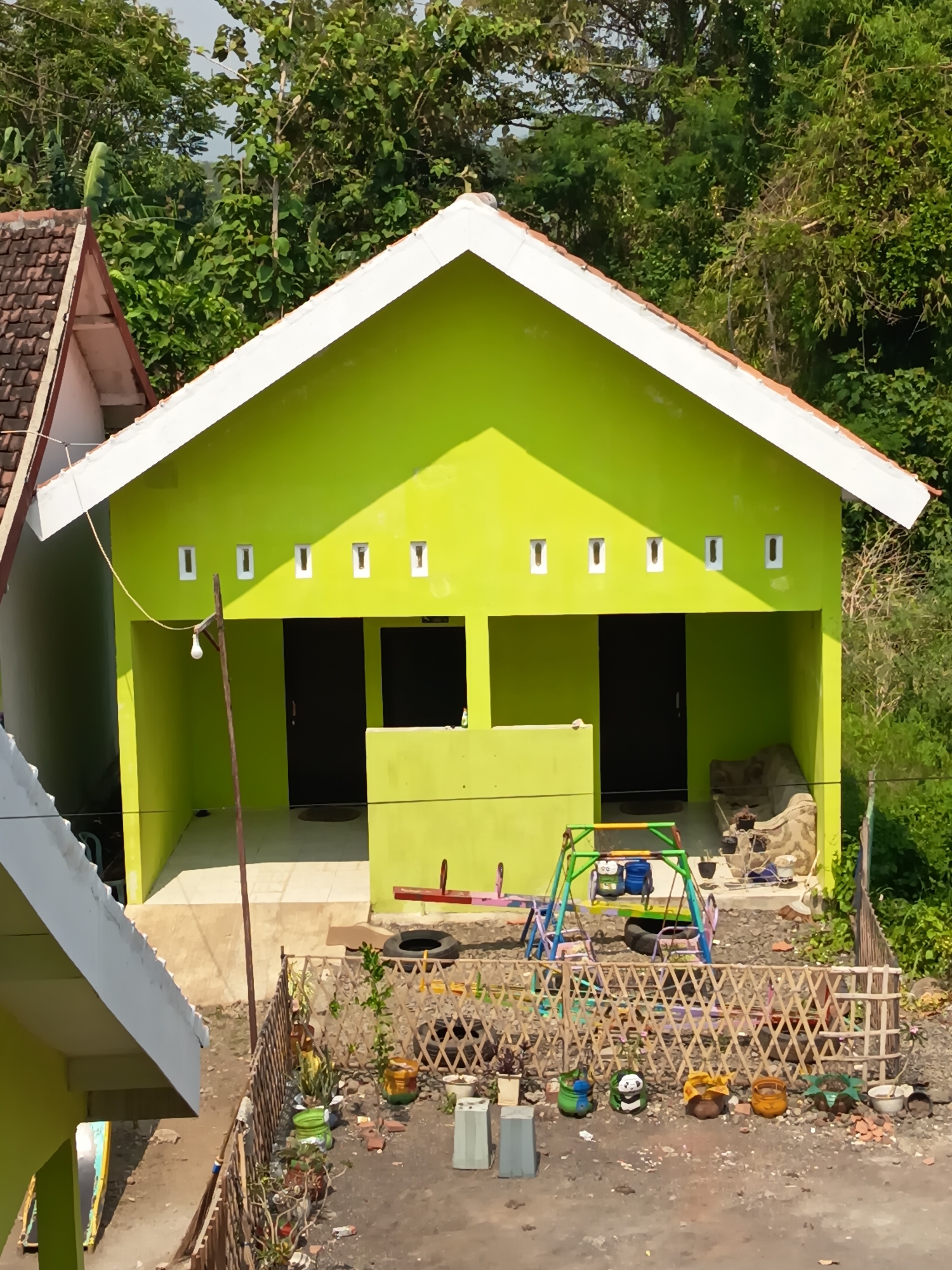 This is a high-angle, eye-level, medium shot of a small, bright green building with a white-trimmed gabled roof, situated in Widoro, Indonesia. The building has two dark-colored doors and a series of small square windows across the top facade. In front of the building is a small playground with a colorful swing set and a seesaw. A rough wooden fence encloses the area, which contains various potted plants, some with decorative designs, and scattered debris. The background is filled with lush green trees and vegetation. The scene appears to be residential or a small community building.