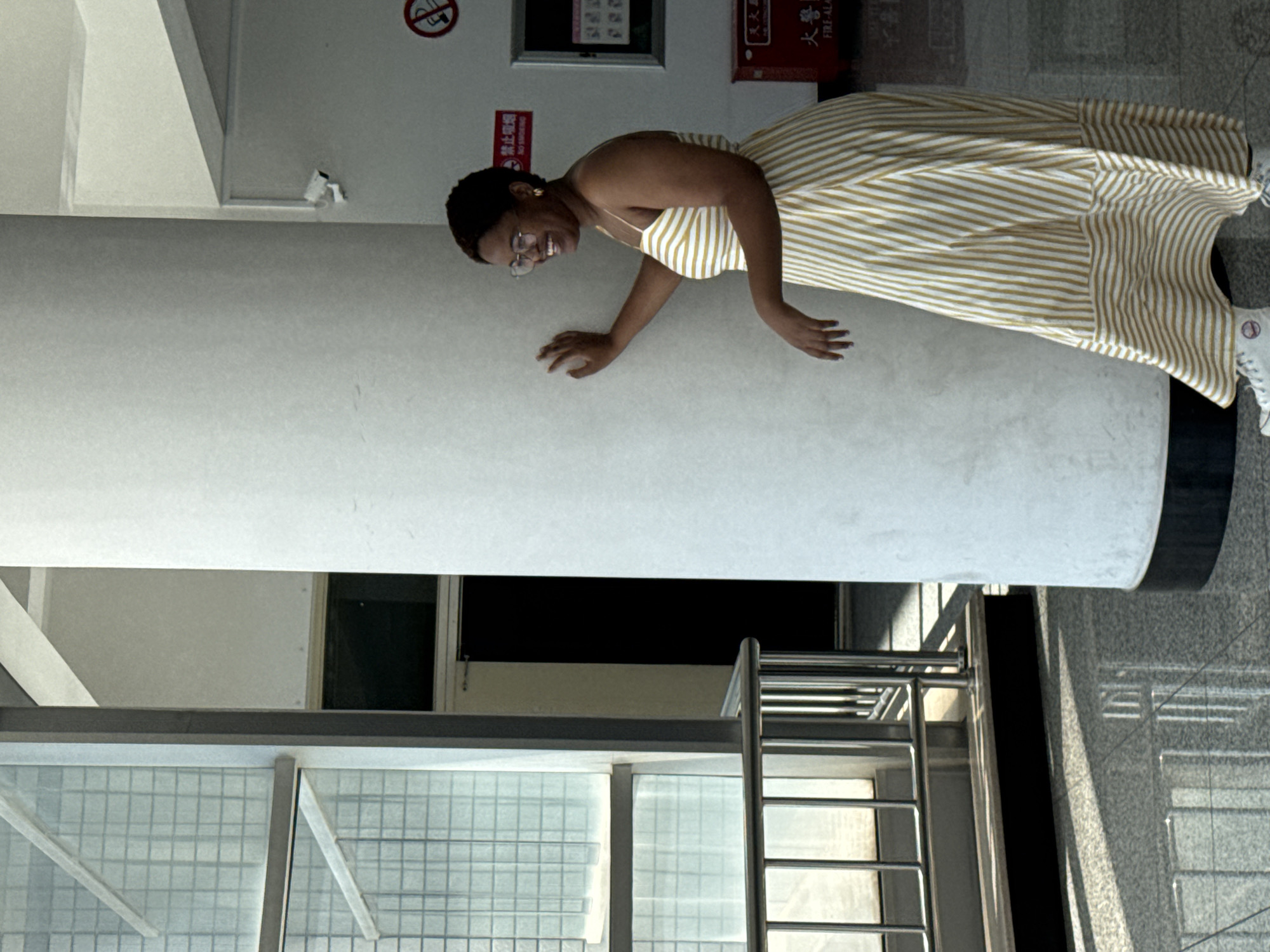 A Black woman stands next to a large, white cylindrical pillar in what appears to be a modern building with polished floors and large windows. She is wearing a yellow and white striped maxi dress with spaghetti straps and white sneakers. Her hair is short and dark, and she is wearing glasses and earrings. She is smiling and looking to her right, with her right hand resting on the pillar.
In the background, there is a large window with a grid pattern, a metal railing, and a dark doorway. A "No Smoking" sign is visible on the wall behind the woman, along with a fire alarm box. The overall lighting suggests an indoor setting with natural light coming from the window. The location is not specifically identified as Guta, China, but the "No Smoking" sign appears to have Chinese characters.