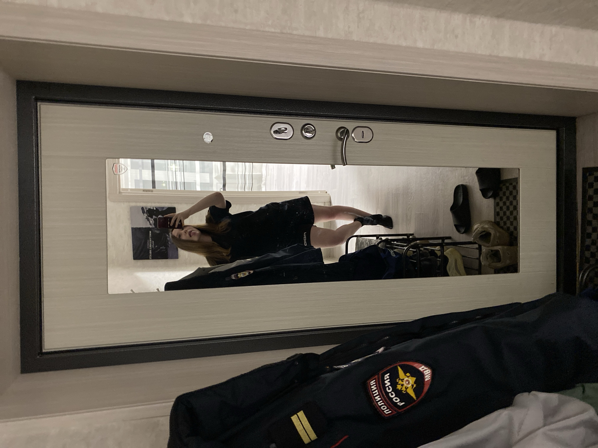 The image shows a mirror reflection of a young woman taking a selfie with her phone. She's wearing a black dress and black shoes. Behind her in the reflection is a rack with various items. In the foreground, outside of the mirror, is a dark blue police jacket with a Russian police patch on it. The background is out of focus, but the reflection shows a window overlooking a city street with tall buildings. A street sign visible in the reflection reads "Чайковский". The location is Chaikovsky, Russia.