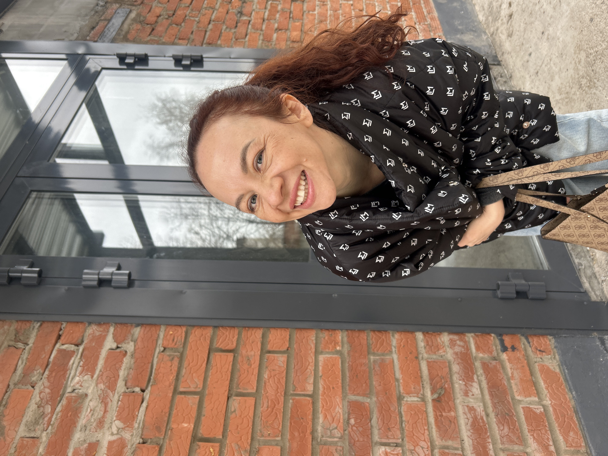 Here's a detailed description of the image:
**Scene:** An outdoor shot featuring a woman posing against a brick wall with a modern-style window structure.
**People:** A smiling woman with reddish-brown hair pulled back in a ponytail. She is wearing a black patterned jacket.
**Environment:** The setting appears to be urban, with a building façade visible. A brick wall with a gray window structure is present. The window reflects a cloudy sky and bare trees.
**Objects:**
* Black patterned jacket
* Window with a modern design
* Brick wall
* Handbag
* Jeans
**Actions/Interactions:** The woman is smiling and posing for the camera.
**Location:** The picture was taken near the Chaikovsky Brick Factory in the city of Chaikovsky, Russia.
**Perspective:** The image is taken from a slightly low angle.