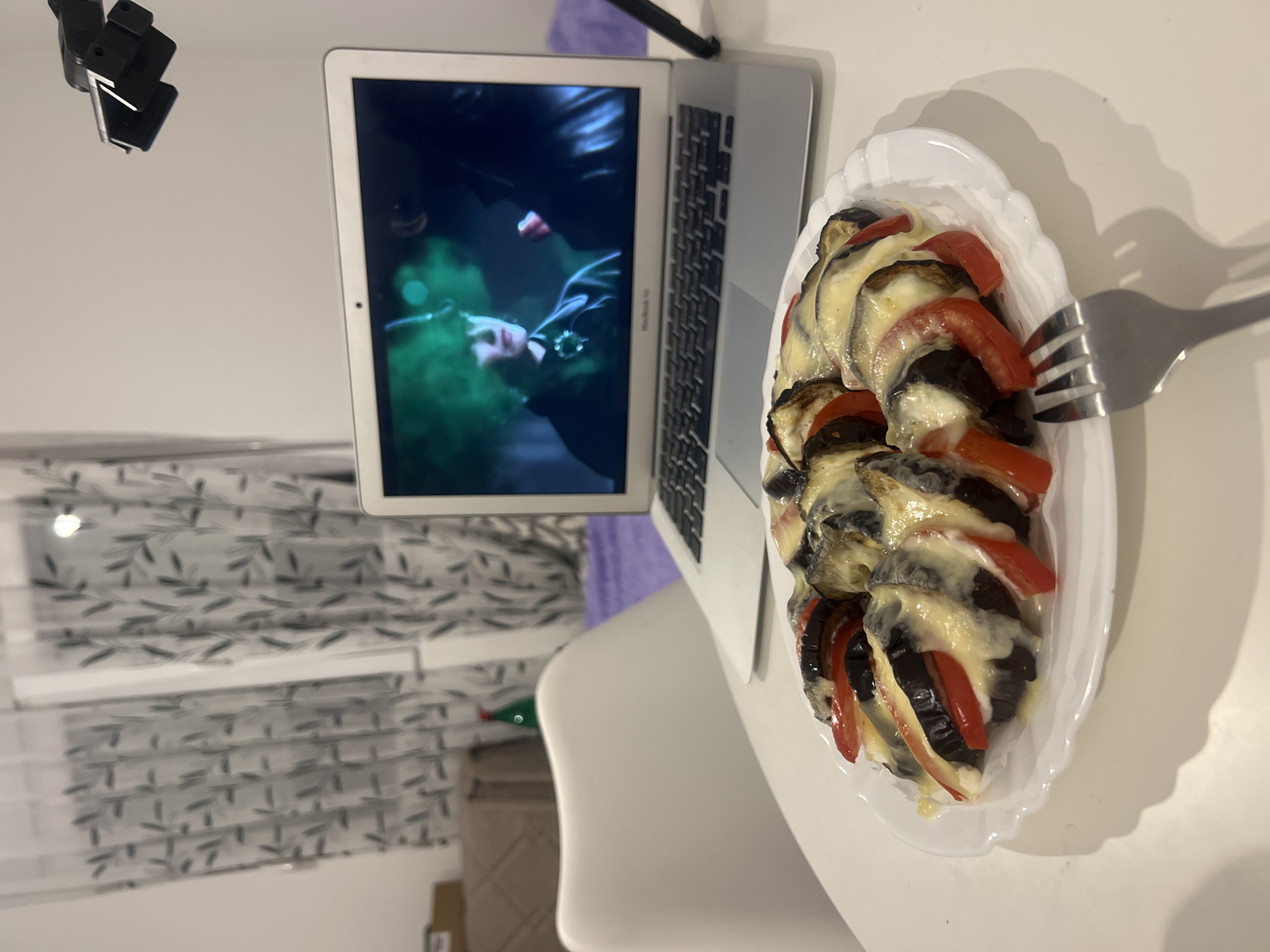 This media file captures an indoor scene, likely a dining or living area, featuring a solitary meal accompanied by digital entertainment. The primary subjects are a white oval plate of food, a laptop, and a fork, all resting on a white table.
The food on the plate appears to be a vegetable dish, possibly a gratin or bake, composed of layered dark-skinned eggplant slices, bright red bell pepper pieces, and melted white or creamy cheese. A silver metal fork is positioned near the plate, casting a distinct shadow on the table, suggesting it has either just been used or is about to be used.
Adjacent to the food, an open silver laptop, identifiable as a MacBook Air (text "MacBook Air" is faintly visible near the hinge), is placed on the table. Its screen displays a still from a video or movie, showing a person in dark clothing within a dimly lit, green-hued scene.
In the background, on the left side, patterned curtains with a grey/green leaf motif are partially visible, suggesting the presence of a window. A blurred brown box or crate and a green bottle are also discernible further in the background. In the upper left corner, a black clip-on device, possibly a phone holder or light, is attached to an unseen object.
The overall scene depicts a relaxed activity of eating a meal while watching something on a laptop. The lighting appears to be artificial, casting clear shadows, which could indicate an evening or nighttime setting indoors. The location context provided is Budva, Montenegro, though no specific visual cues in the image directly identify this location.